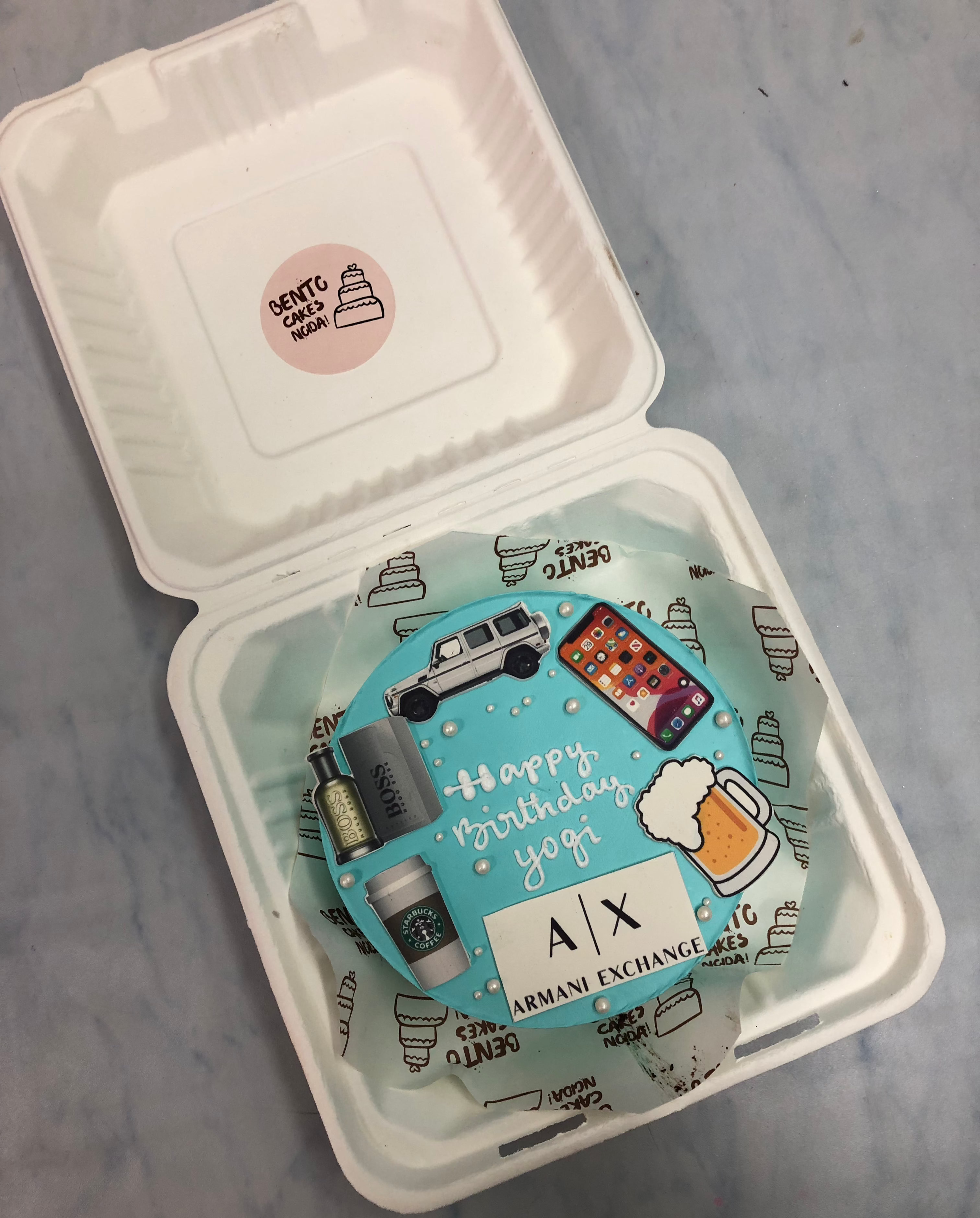 A blue-iced bento cake inside a white container, decorated with various elements like a car, a beer mug, a calculator, Starbucks cups, and an Armani Exchange logo. The cake features "Happy Birthday Yogi" written on top, celebrating a special occasion.