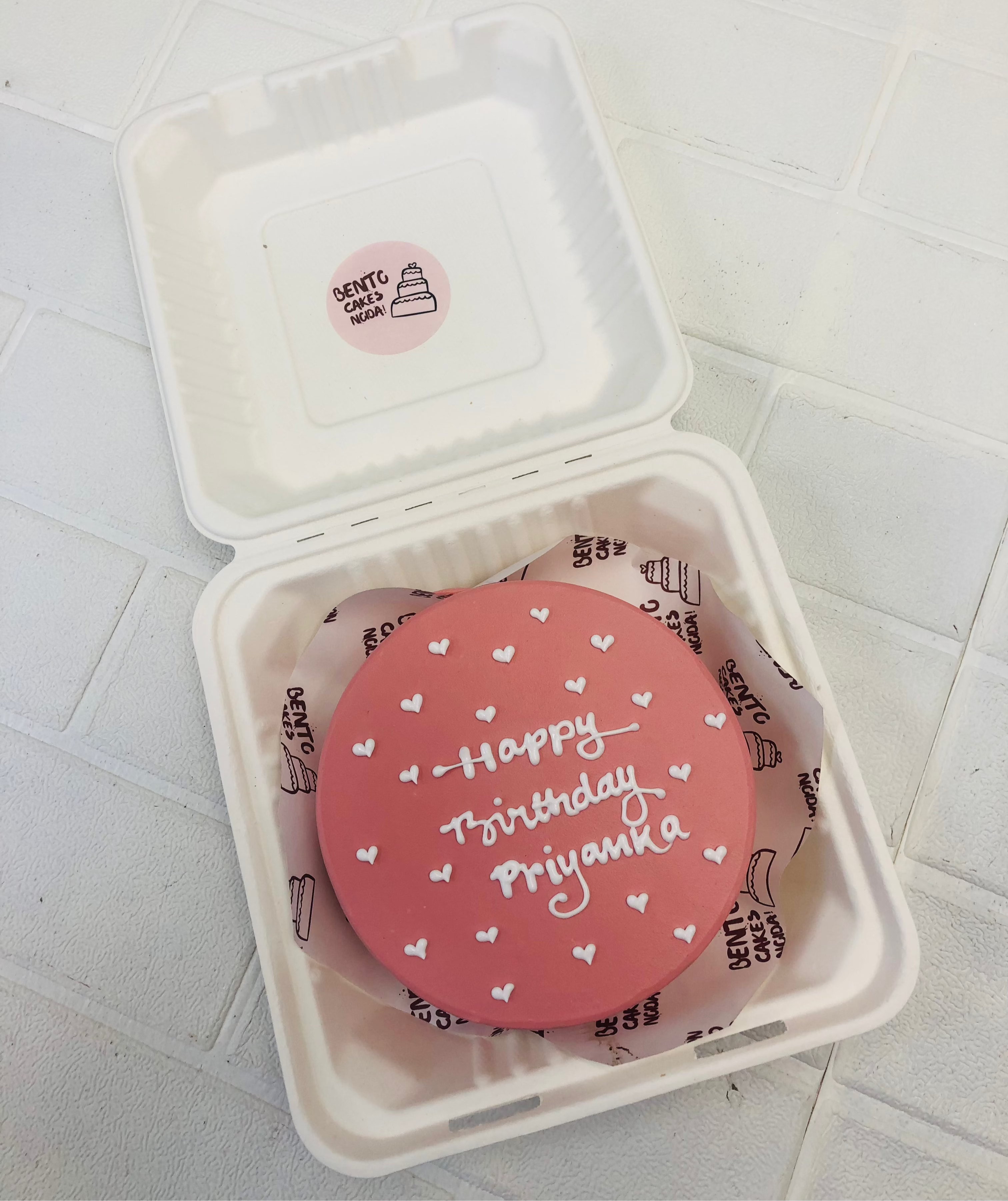 A magenta bento cake decorated with little white hearts and a text "" Happy Birthday Priyanka" written with white icing.