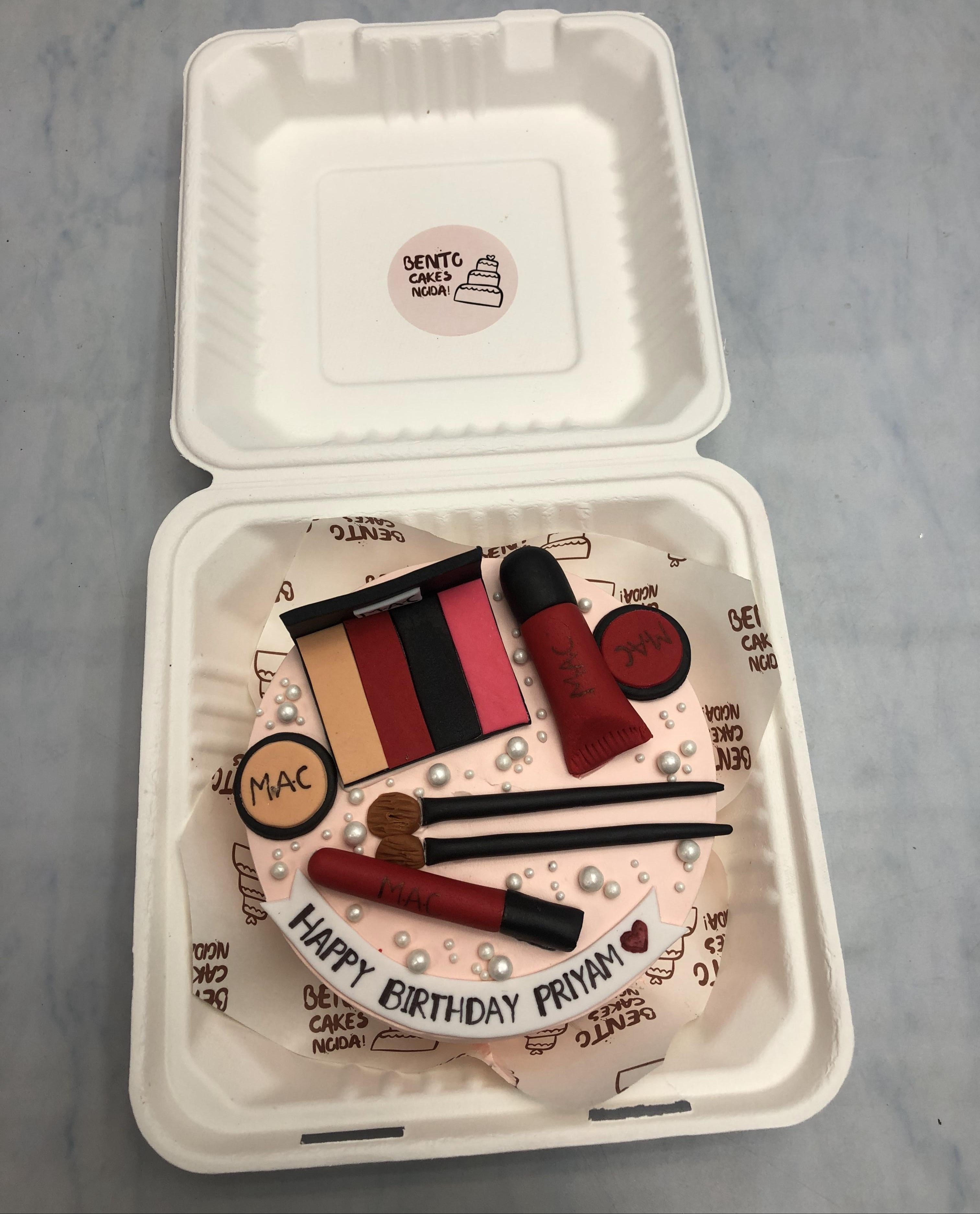 A creatively crafted makeup-themed birthday cake in a white bento box. The cake is adorned with fondant makeup items, including a MAC compact, eyeshadow palette, lipstick, lip gloss, and makeup brushes. White pearls add an elegant touch, and the birthday message "HAPPY BIRTHDAY PRIYAM" is displayed on a ribbon at the bottom.