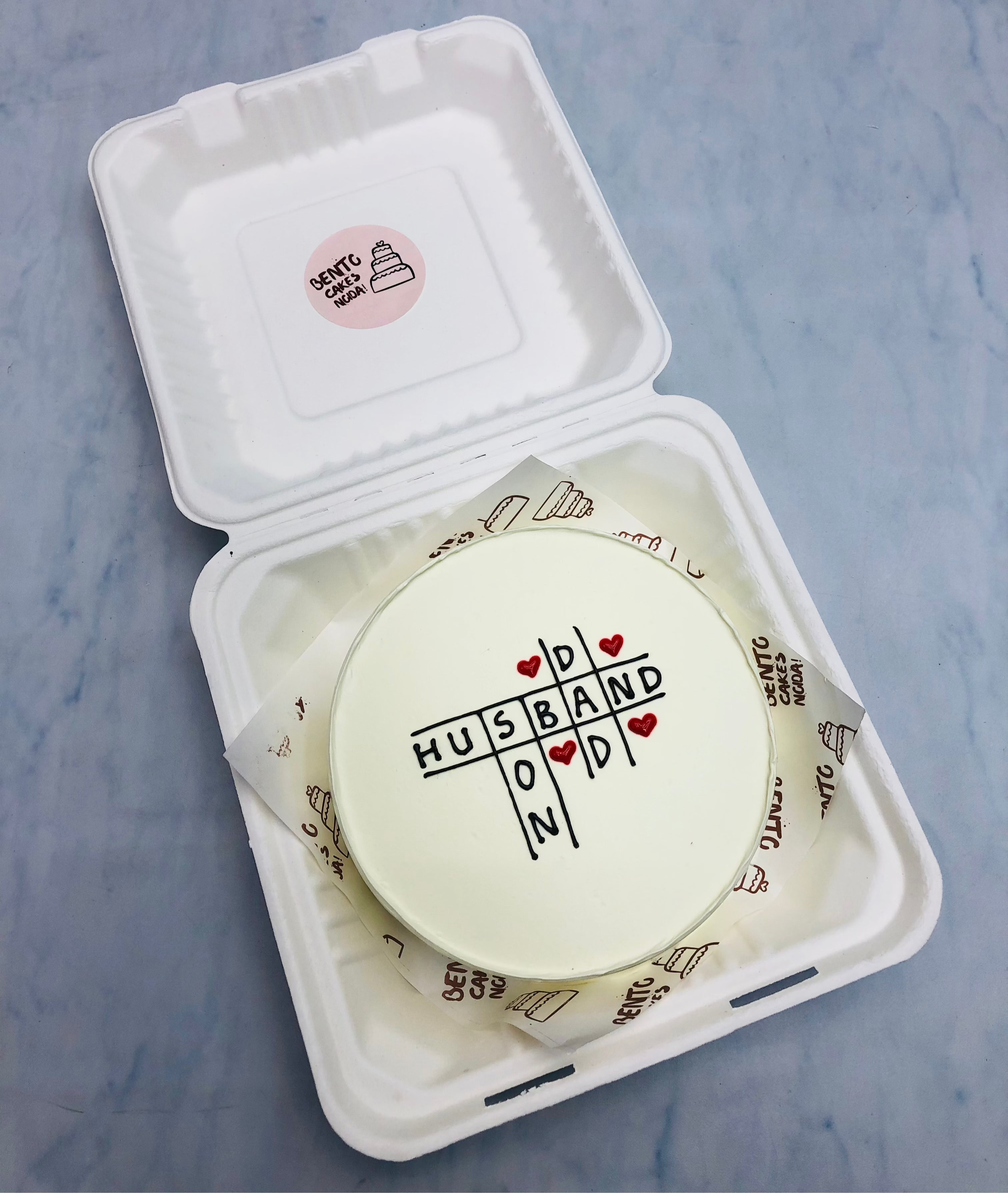 A round cake featuring a crossword puzzle design with the words "HUSBAND" and "DAD" intersecting, decorated with red hearts. The cake is placed inside an eco-friendly white container branded with "CETTU CAKE."