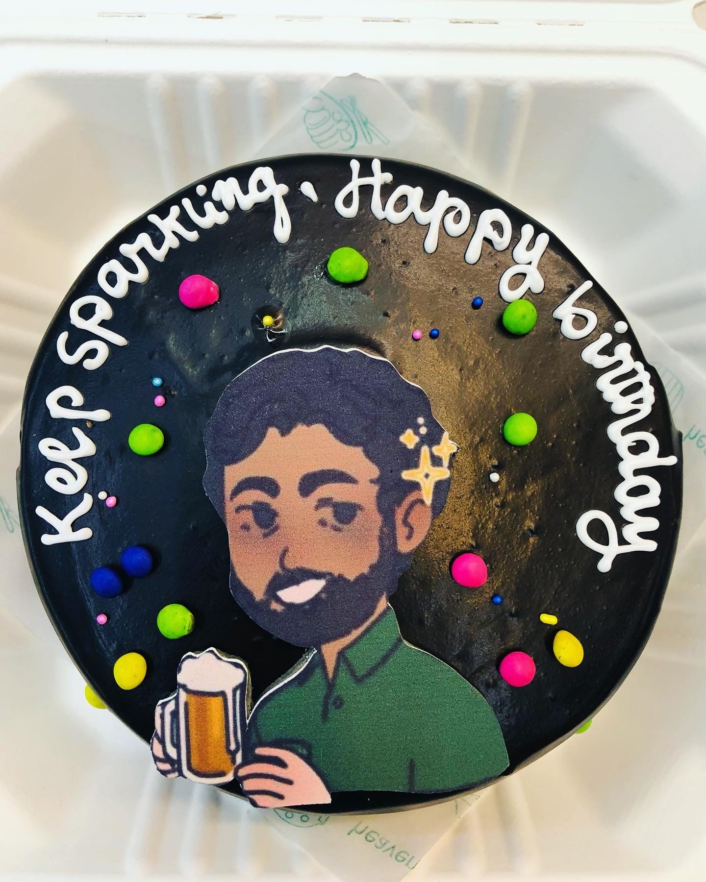 A round black birthday cake with colorful decorations and a message written in white icing that says "Keep sparkling, Happy birthday." The cake features a cartoon image of a person holding a beer mug.