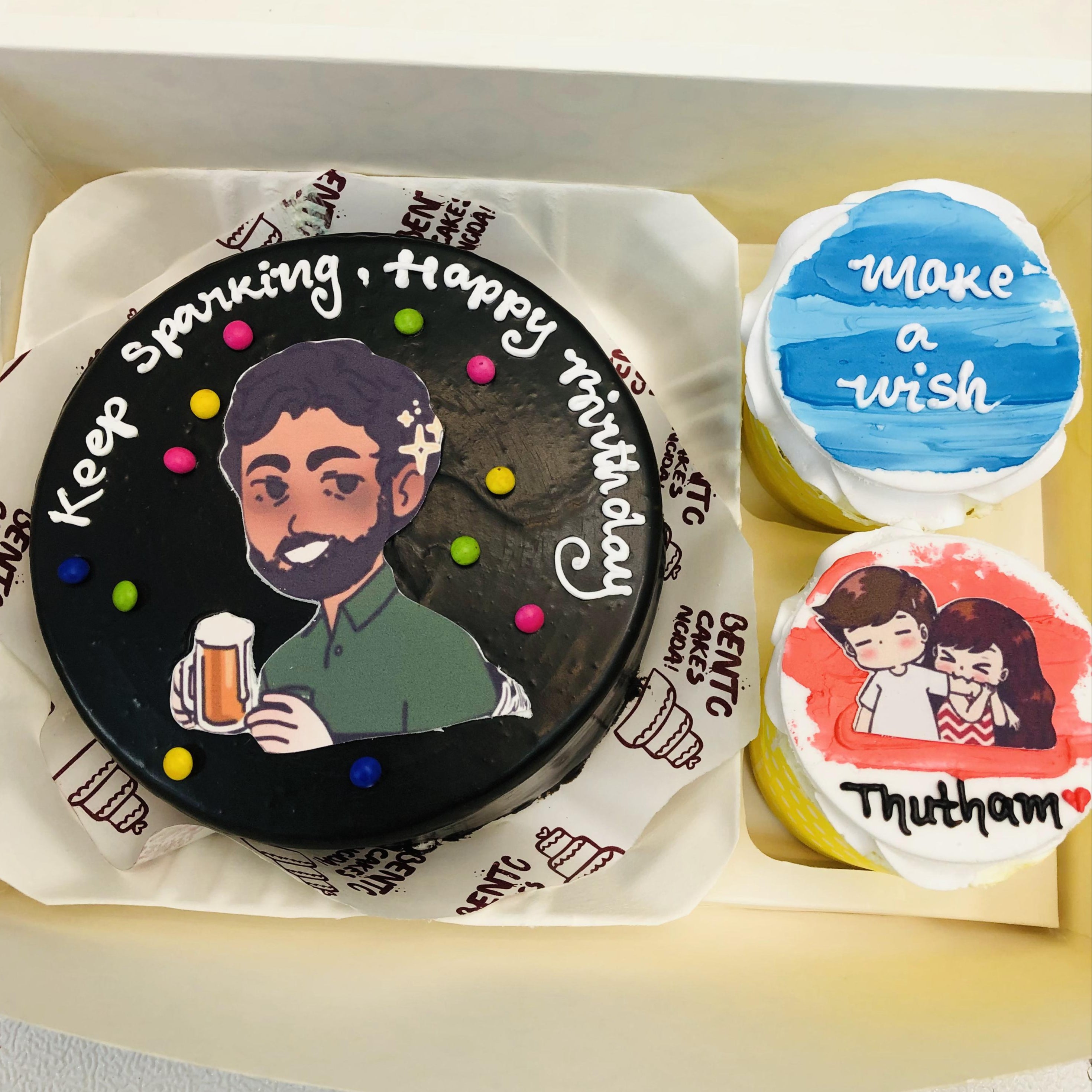 A Men's Birthday Mini Bento Combo Cake set, featuring a black main cake with colorful dots, a person illustration holding a beer mug, and the message "Keep sparking. Happy birthday." The set includes two cupcakes: one with "make a wish" in blue and white, and another with a couple's illustration and "Thutham" with a heart symbol.