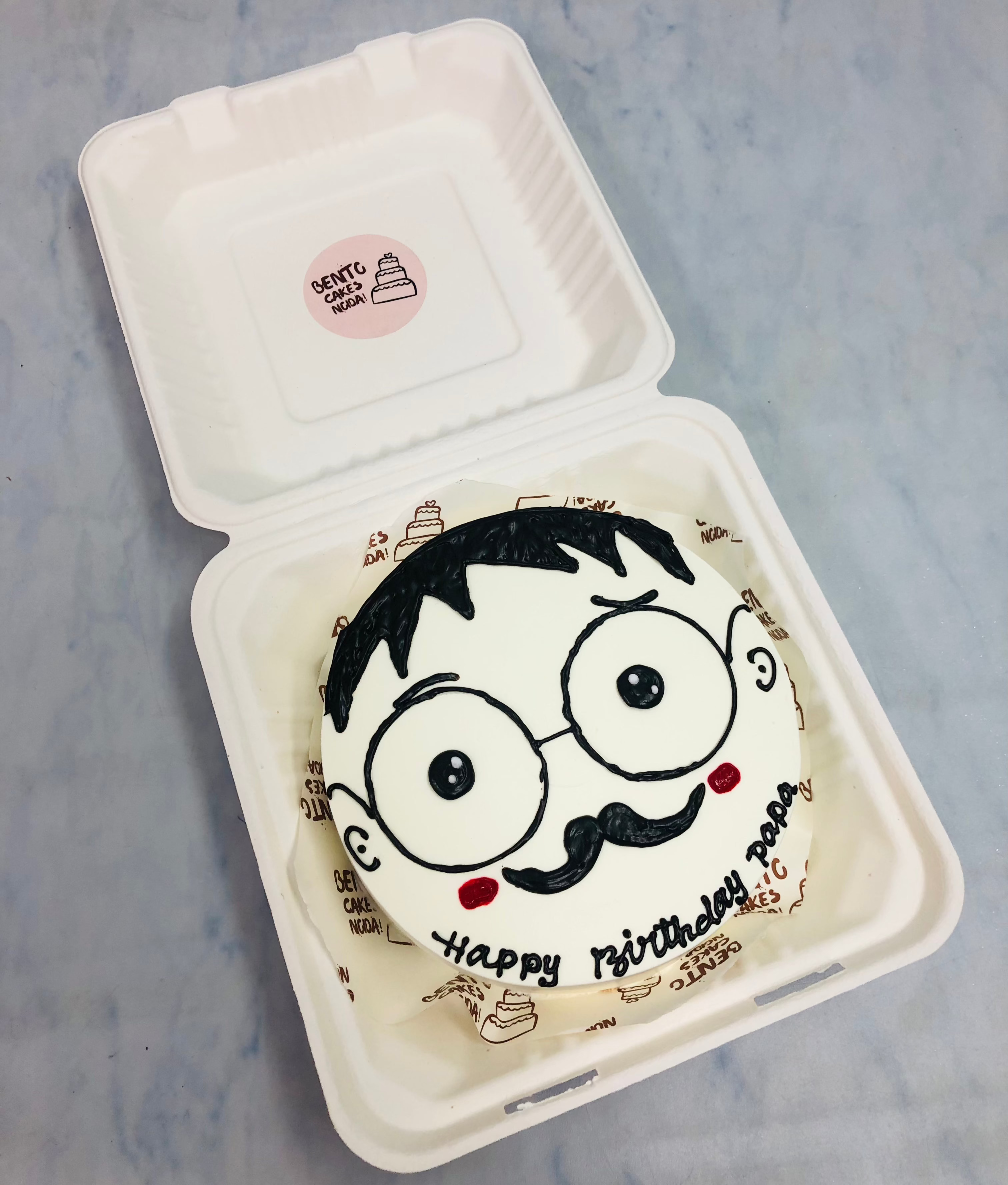 A beautifully designed round birthday cake with a cartoon-style face, including glasses, a mustache, and rosy cheeks. The cake features the text "Happy Birthday Papa" and is placed in a white cake box with a "GENTLE CAKE" sticker on the lid.