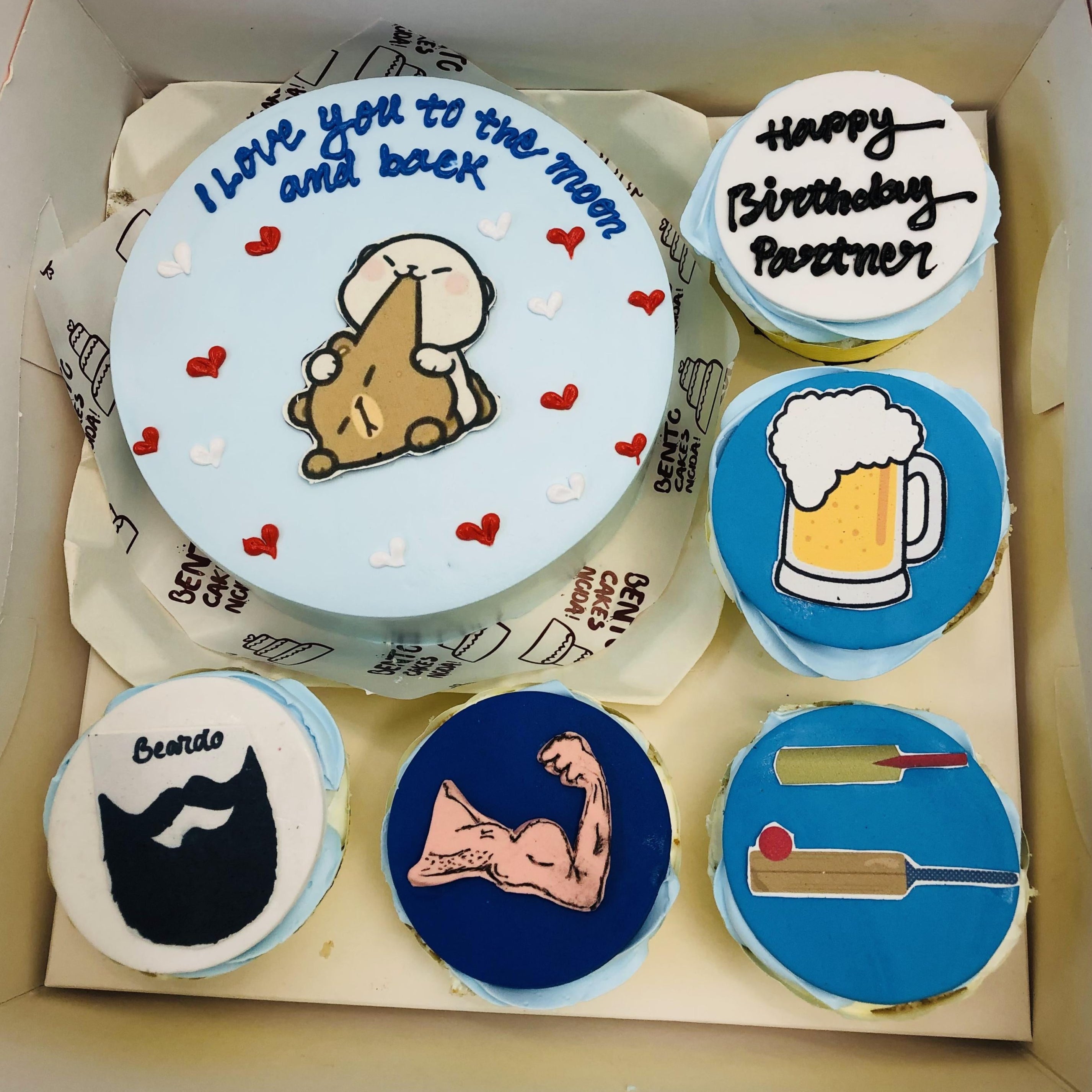 A men's theme bento cake set featuring a large round cake with a character hugging a moon and the message 'I love you to the moon and back,' surrounded by five cupcakes decorated with designs like a beer mug, a muscular arm, a cricket bat and ball, and a beard labeled 'Beardo.'
