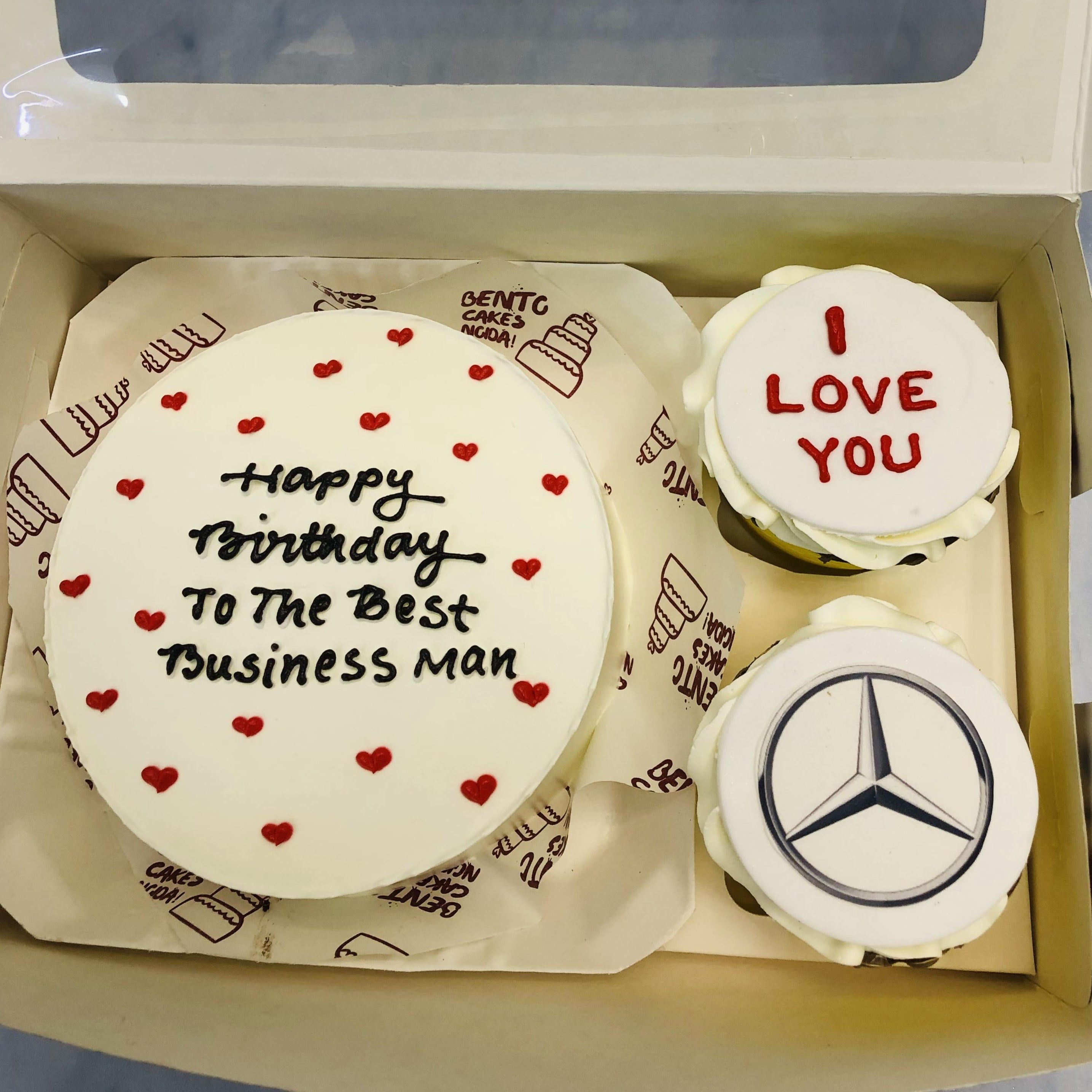 A boxed cake set featuring a large round cake with white frosting, red heart decorations, and the message "Happy Birthday To The Best Business Man" in black icing. Accompanied by two smaller cakes: one with the Mercedes-Benz logo and another with "I LOVE YOU" in red icing. The cakes are placed on a paper liner with "BENTO CAKES NIGDA!" written on it.