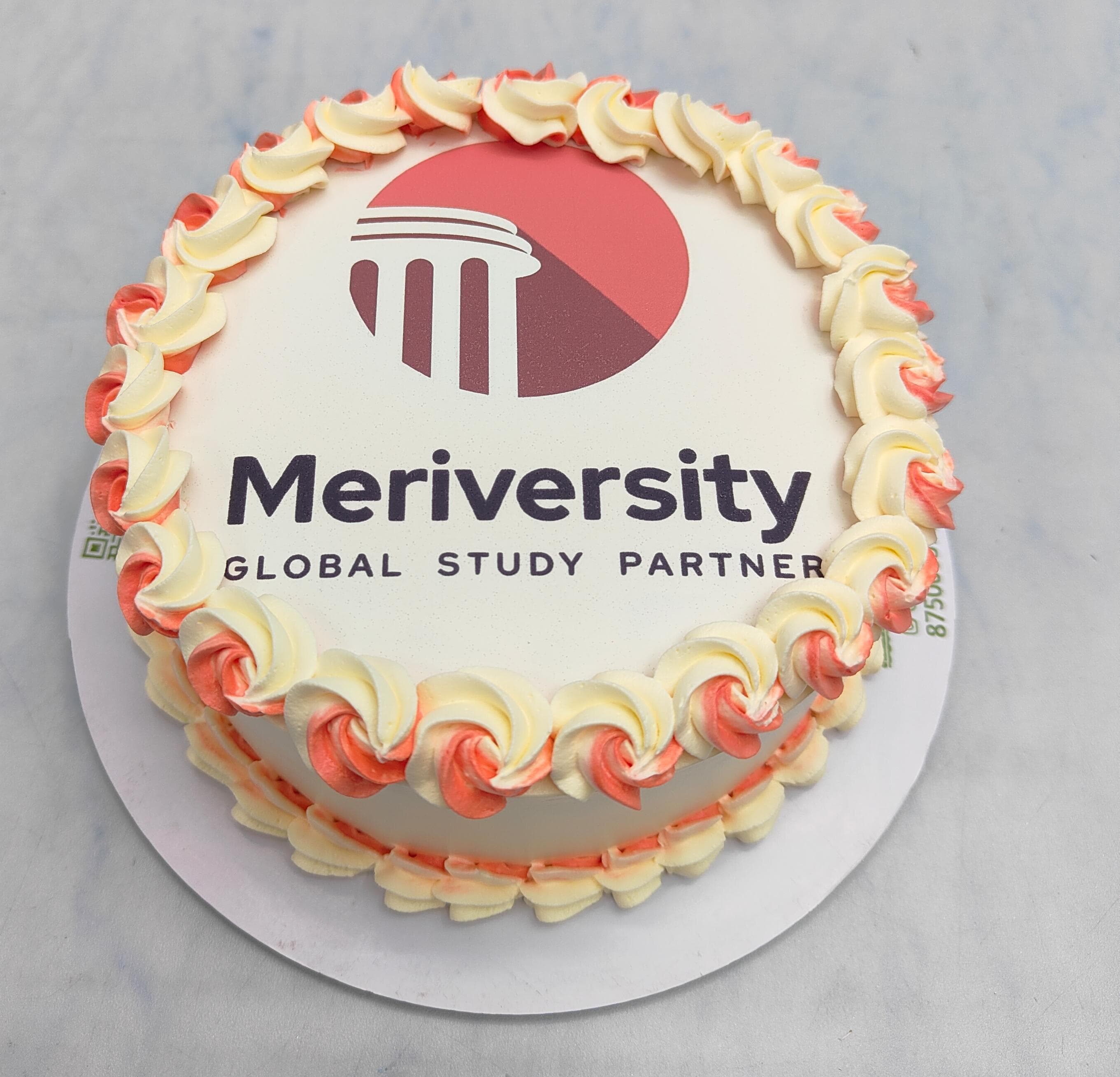 A round cake with elegant white icing, featuring the Meriversity logo and the words "Global Study Partner," framed by red and white decorative frosting.