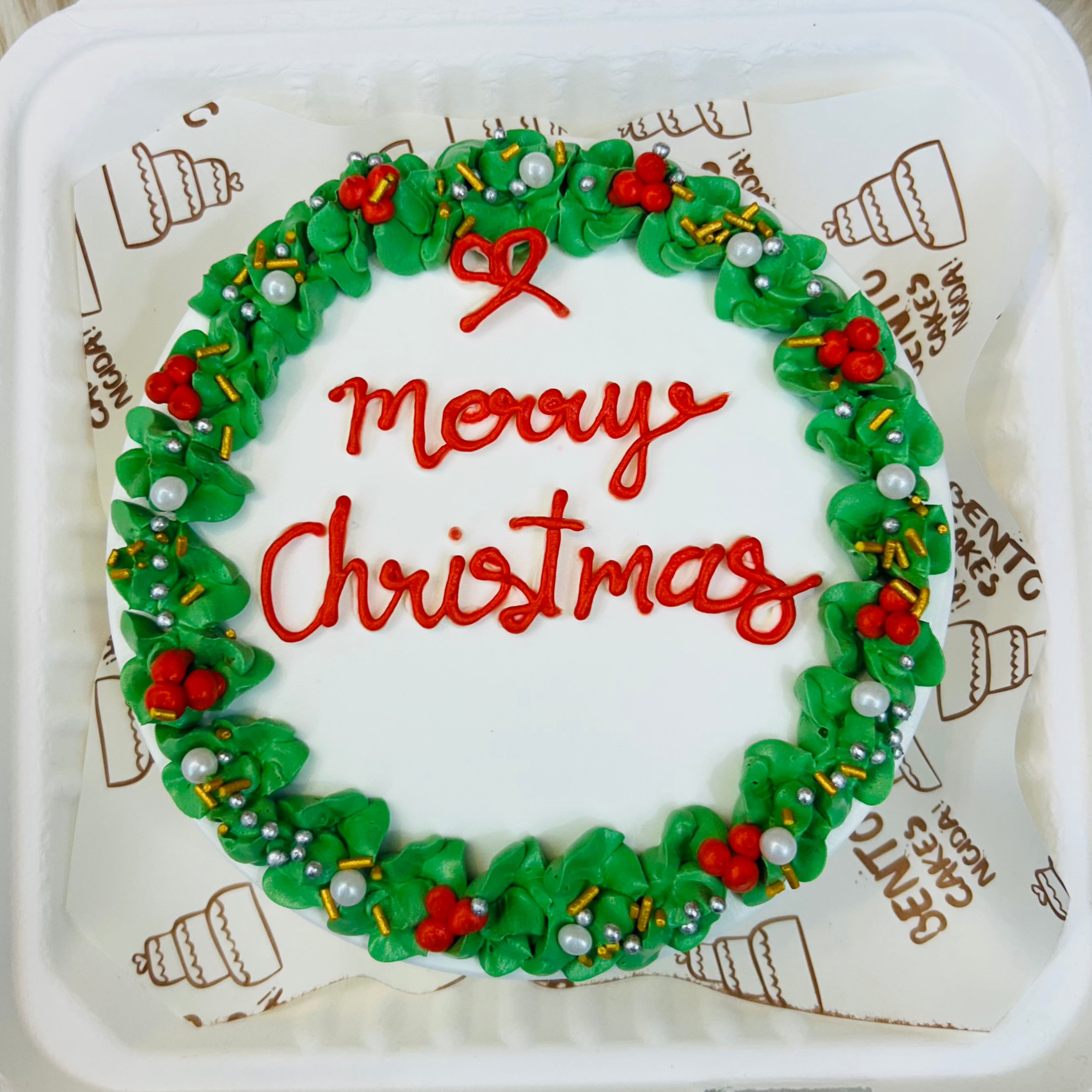 A white bento cake with green florals design on its border with a text in red color "Merry Christmas" in the center.