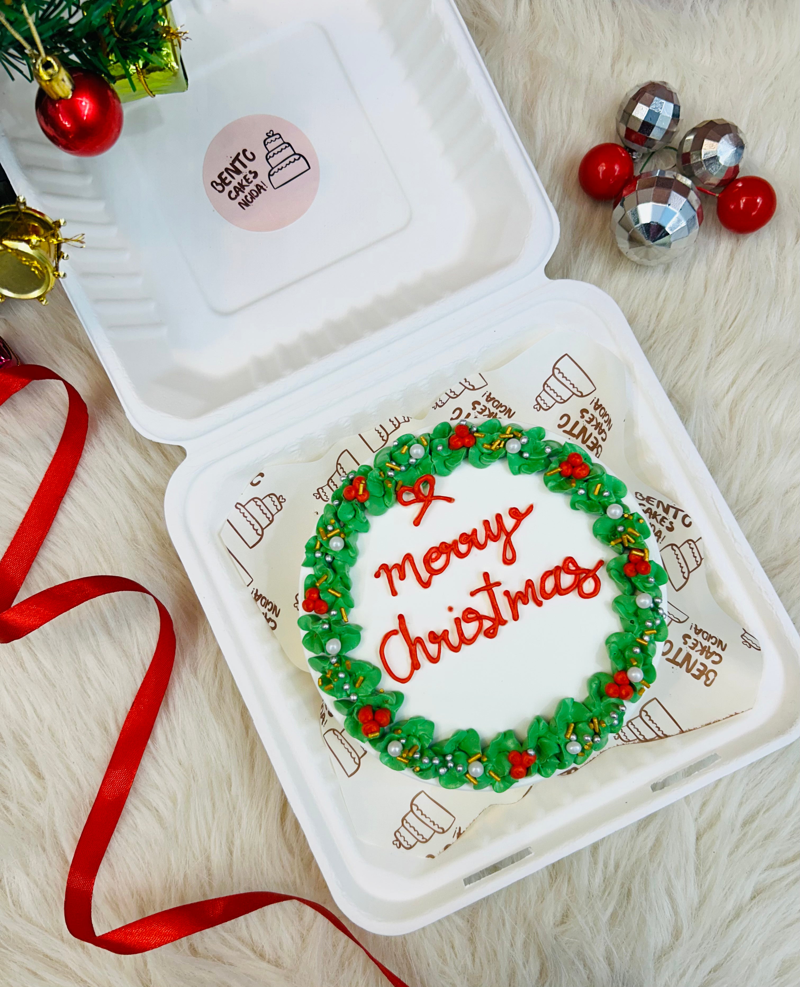 A white bento cake with green florals design on its border with a text in red color "Merry Christmas" in the center.