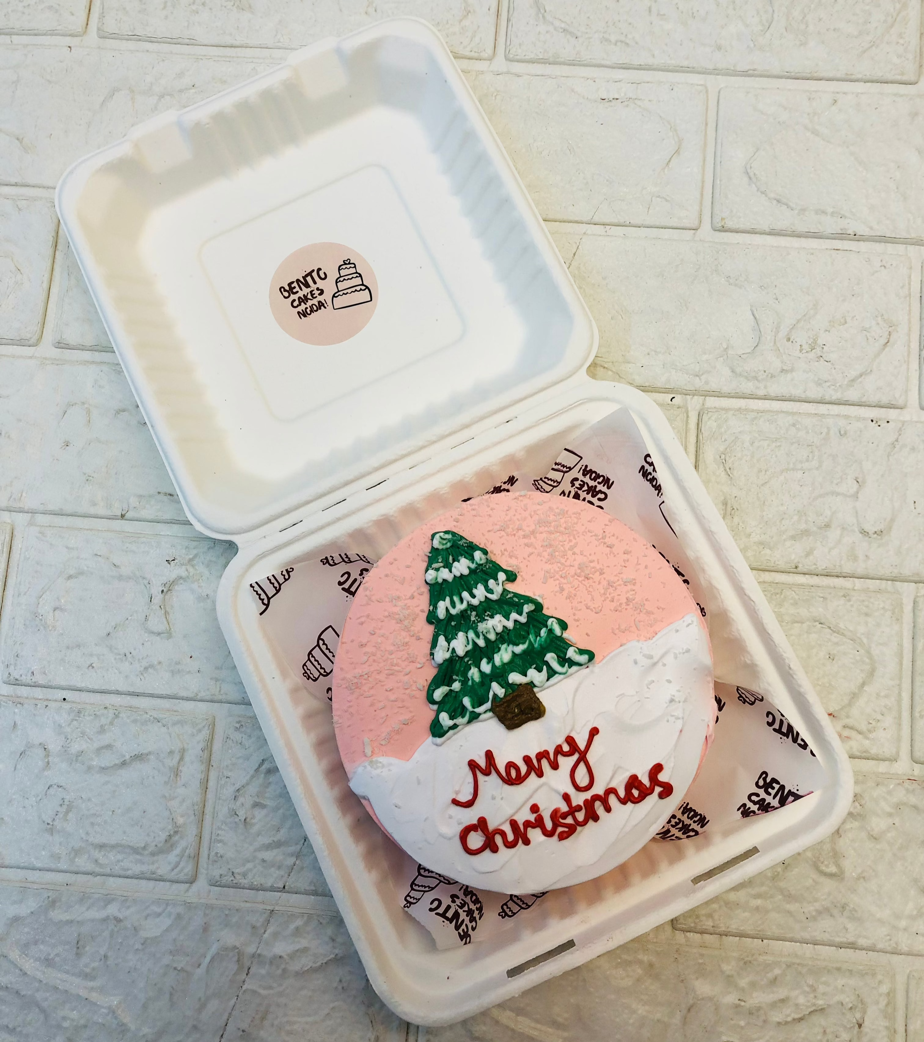 A rose gold and white mix mento cake is designed with a christmass tree on top with a Merry Christmas Text.