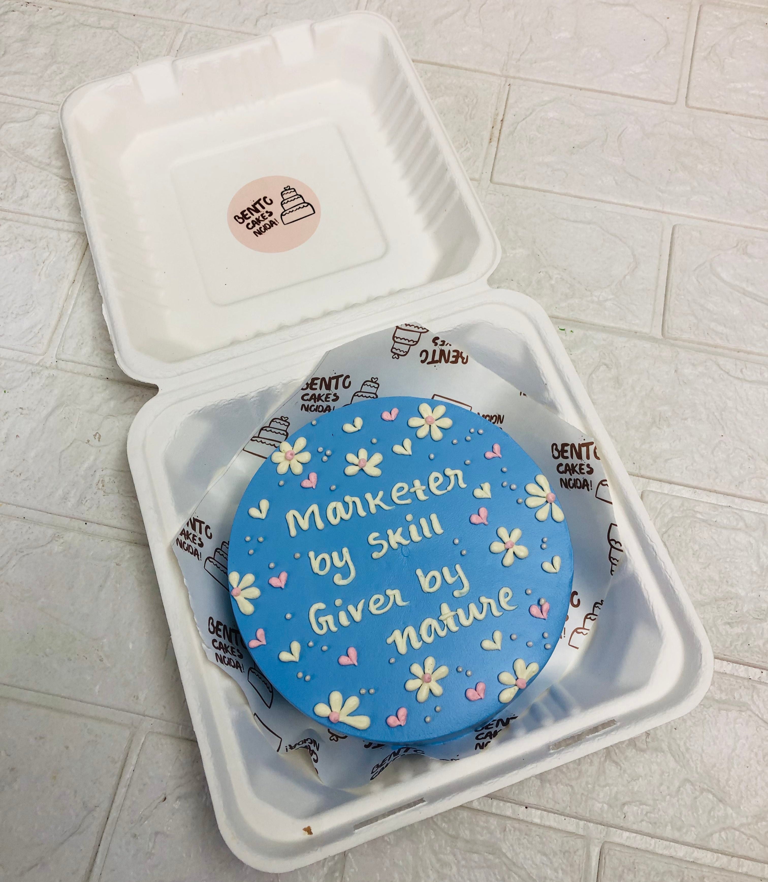 A beautifully decorated blue bento cake with delicate white and yellow flower accents. The cake features an uplifting message: "Marketer by skill, Giver by nature," written in white icing.