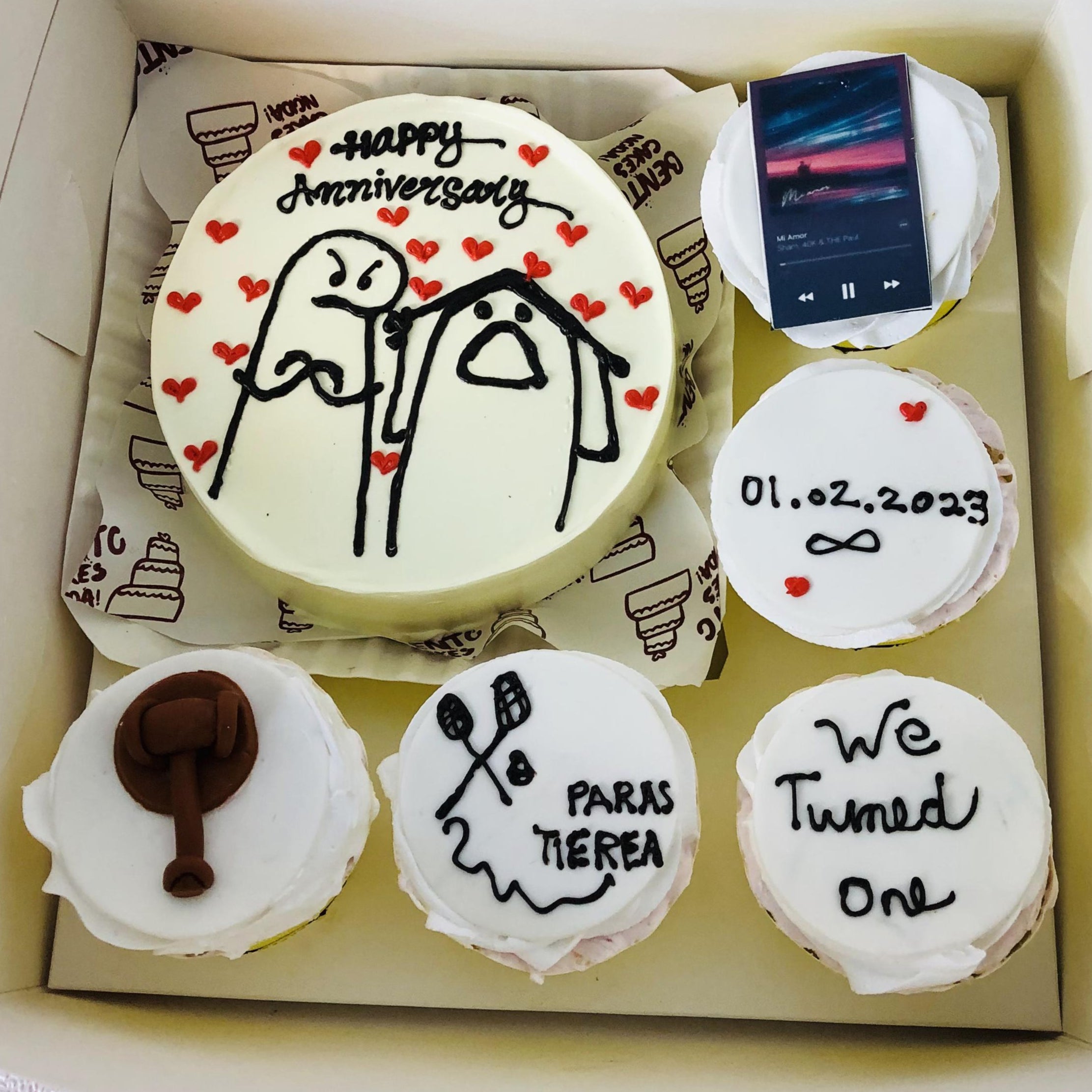 A bento combo cake set featuring a large round cake with cartoon characters surrounded by hearts and the text 'Happy Anniversary,' accompanied by four unique cupcakes with decorations including a song player, an anniversary date, a gavel, and crossed paddles.