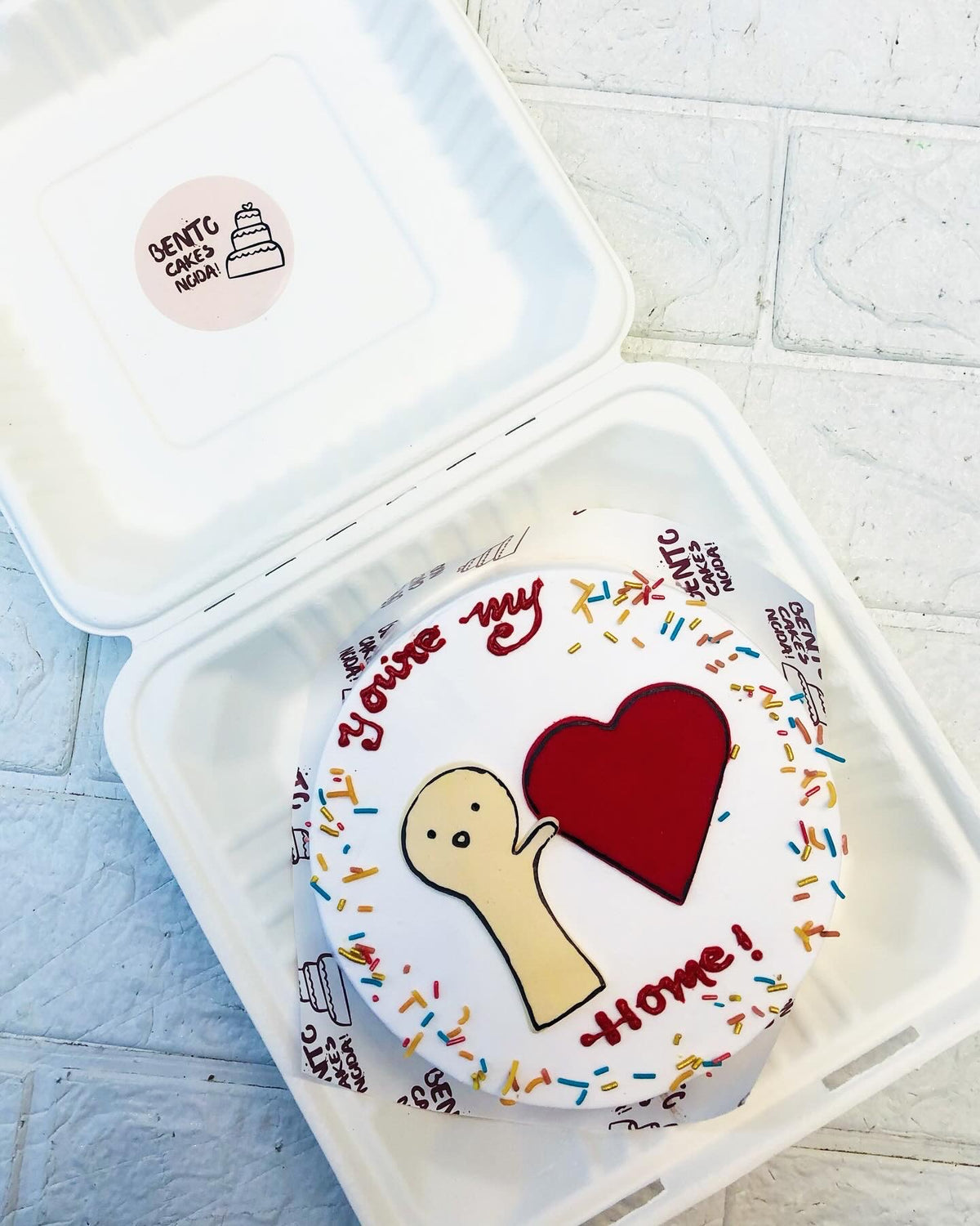 A beautifully decorated bento cake in a white container, featuring a cartoon figure holding a red heart. The cake is adorned with colorful sprinkles and the message "You're my Home!" in red icing, making it a perfect gift to express love and warmth.