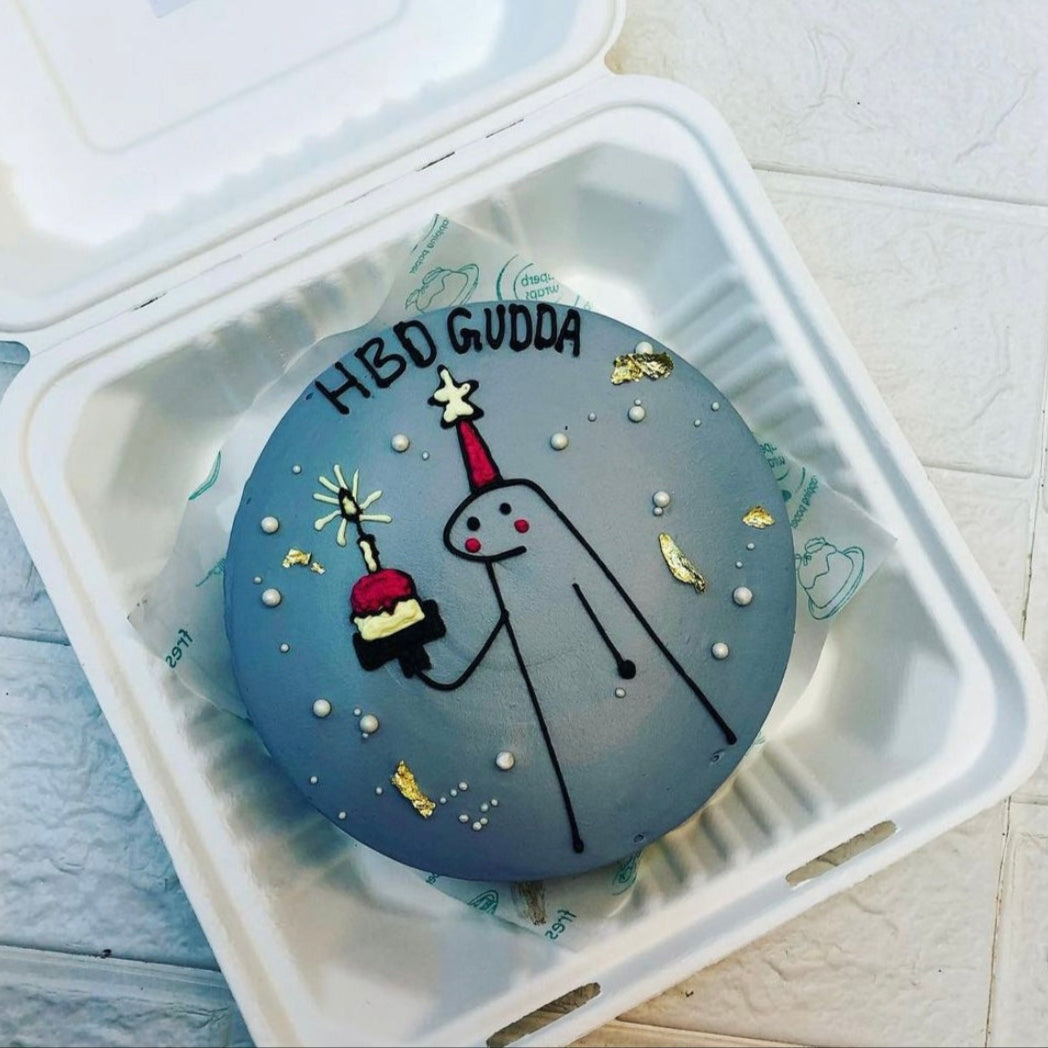 A round birthday cake with a gray frosting, featuring a simple drawing of a figure wearing a red party hat and holding a small cake with a lit candle. The cake is decorated with white pearls and gold leaf accents, and the text "HBD GUDA" is written on top. The cake is placed inside a bento box.