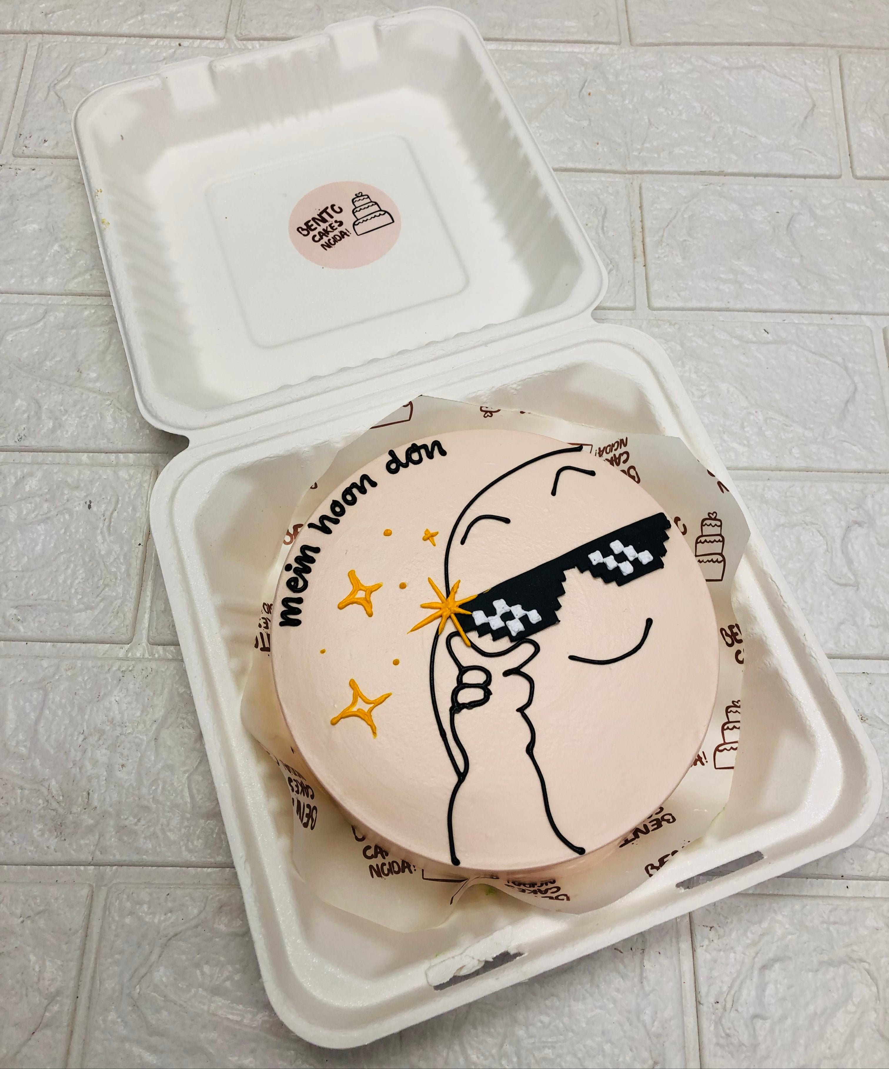 A peach color bento cake having a minimalist cartoon wearing goggles and cake descorated with golden stars.