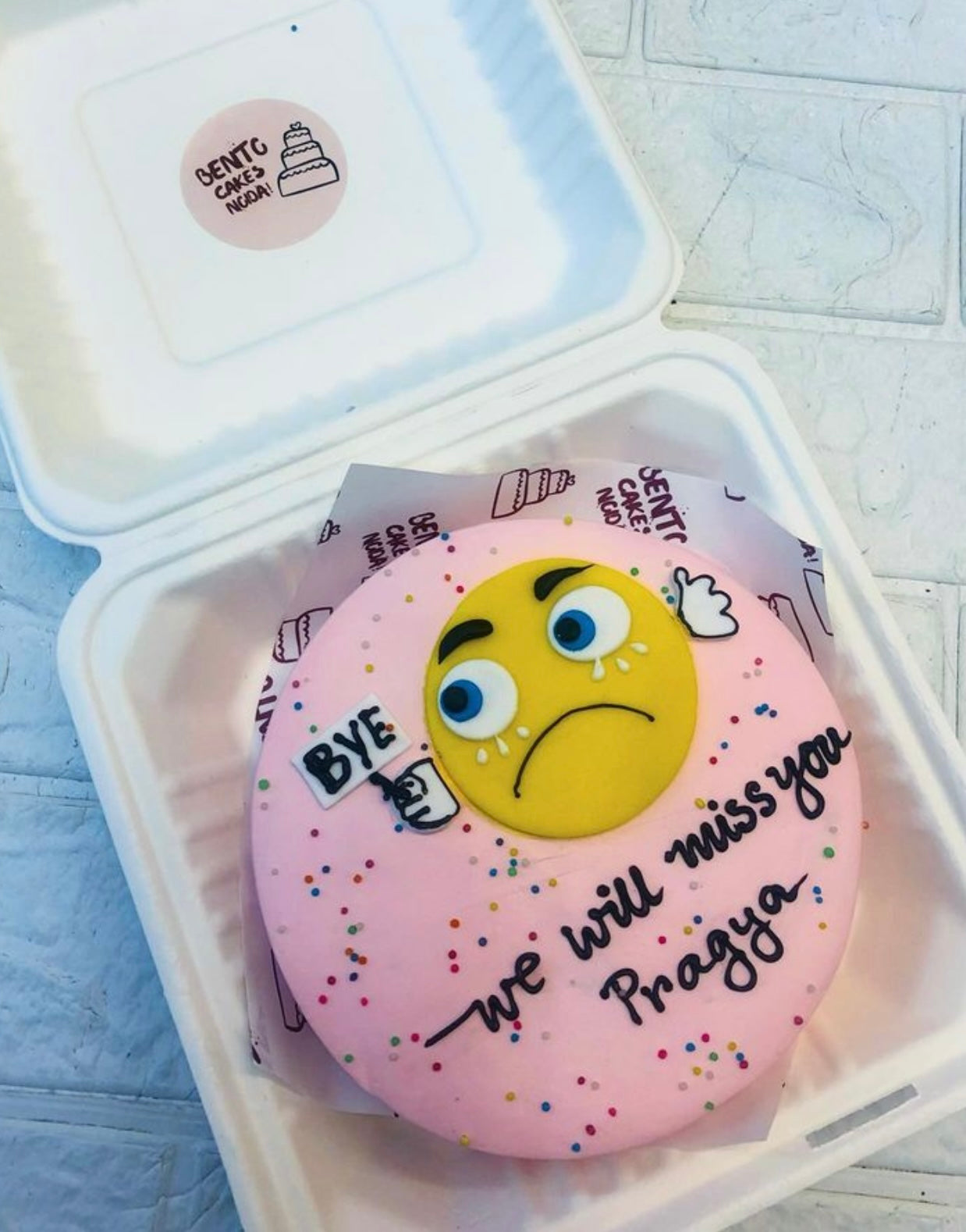 A pink bento cake having a fondant weeping emoji design with a missing text.