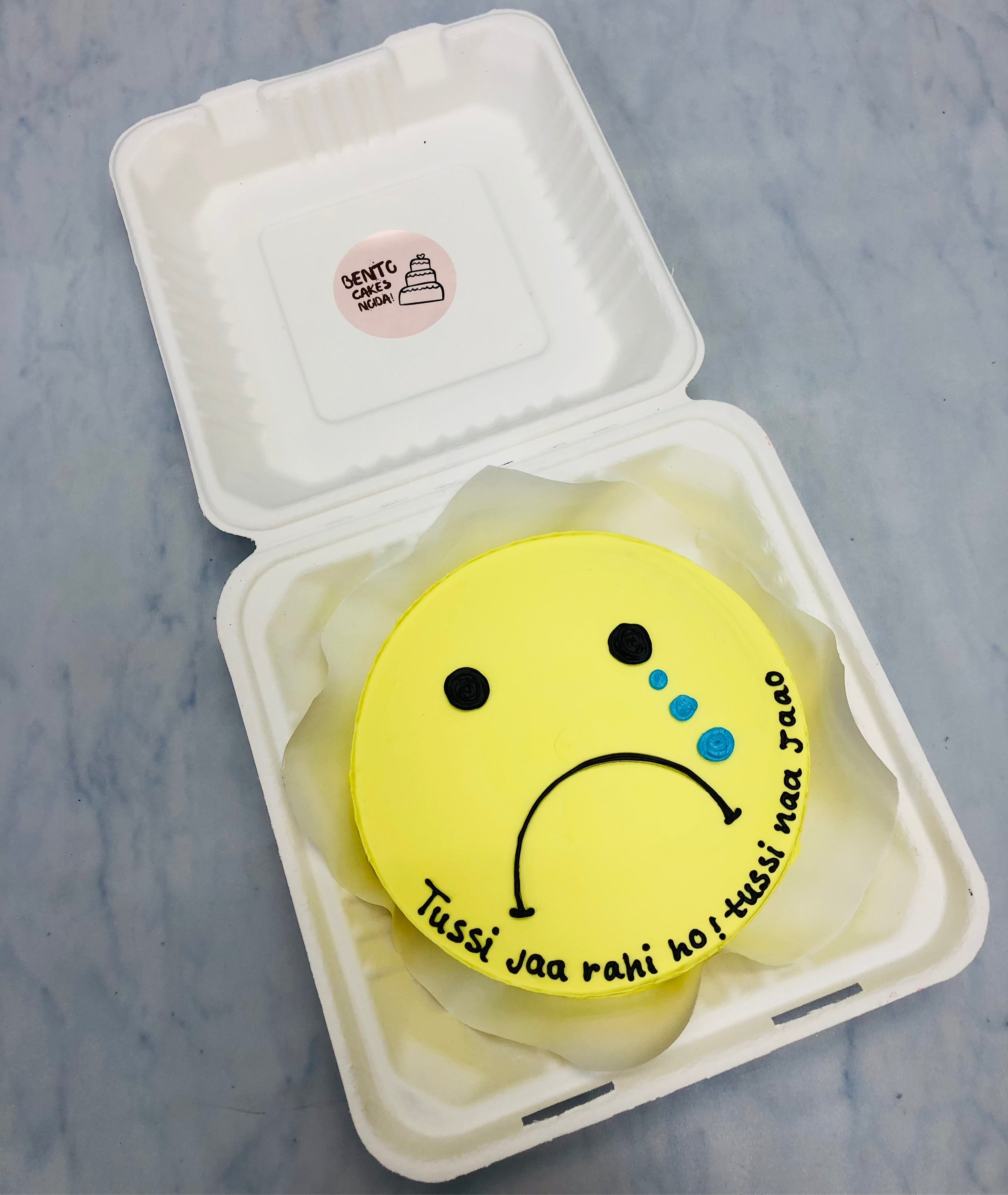 A yellow bento cake is designed for missing person theme with a weeping emoji face on it.
