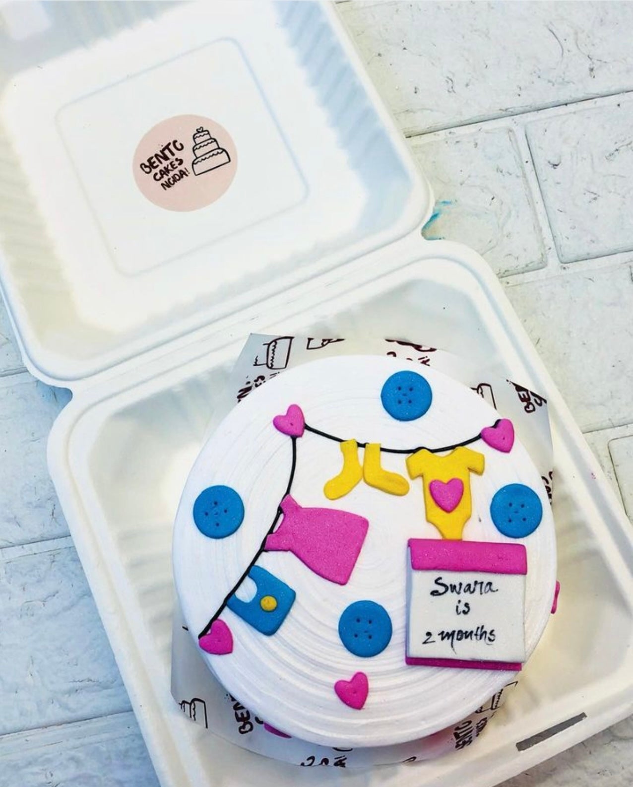 A small round cake with white frosting, decorated with fondant baby clothes and accessories, placed inside a white biodegradable box. The cake features blue buttons, a yellow onesie, a blue bib, and a red and white card with 'Swara is 2 Months' written on it.