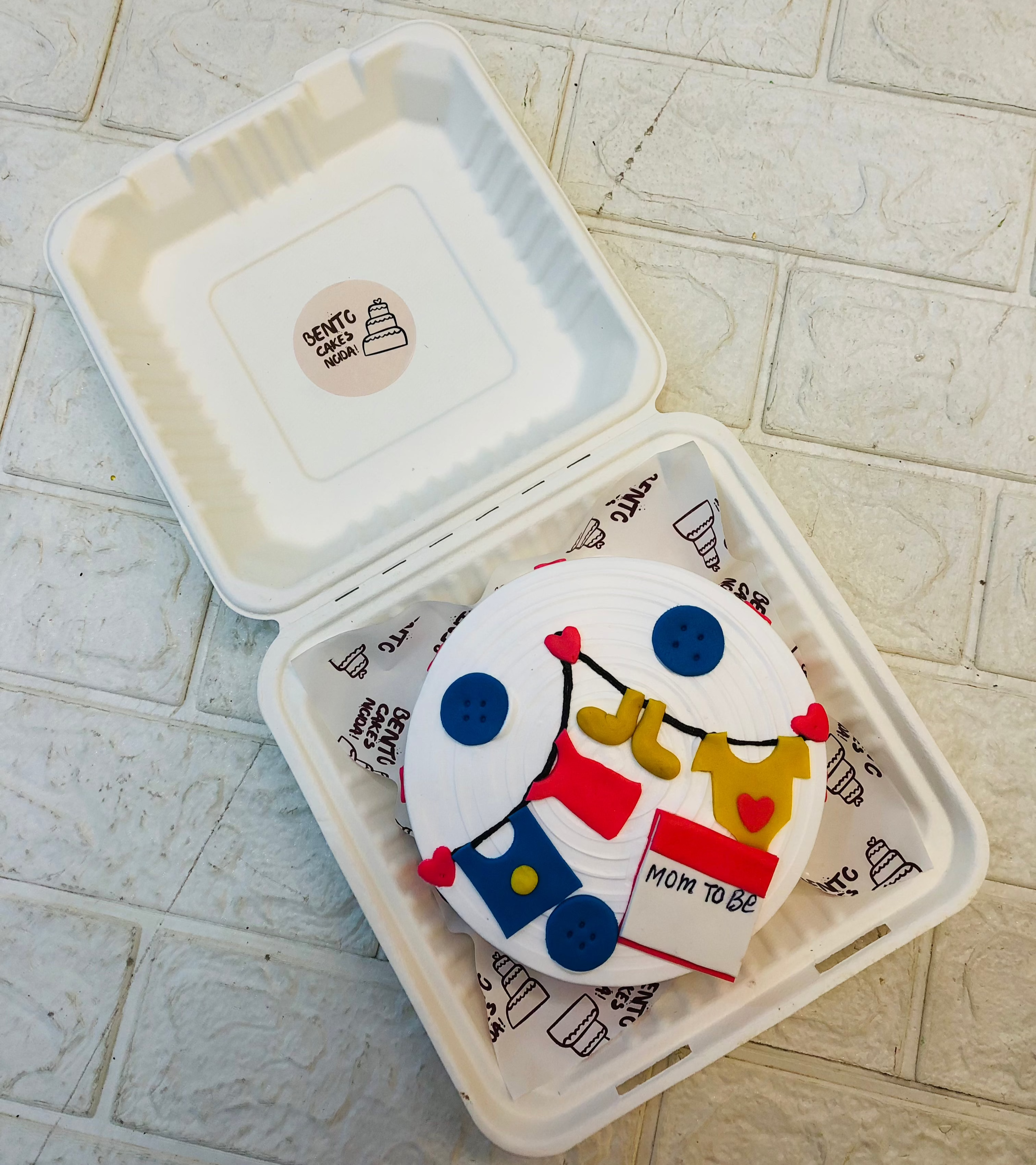 A small round cake with white frosting, decorated with fondant baby clothes and accessories, placed inside a white biodegradable box. The cake features blue buttons, a yellow onesie, a blue bib, and a red and white card with 'Mom to Be' written on it.