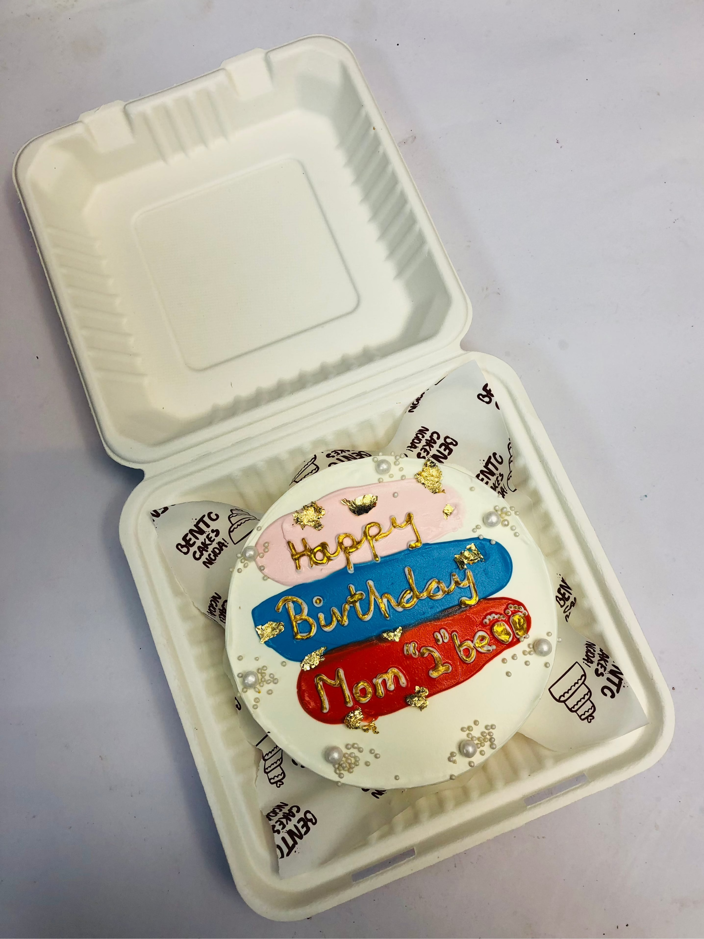 A small round birthday cake placed inside a white biodegradable container. The cake is decorated with colorful icing and gold accents. The icing reads 'Happy Birthday Mom 2 be' in gold letters on pink, blue, and red backgrounds. 