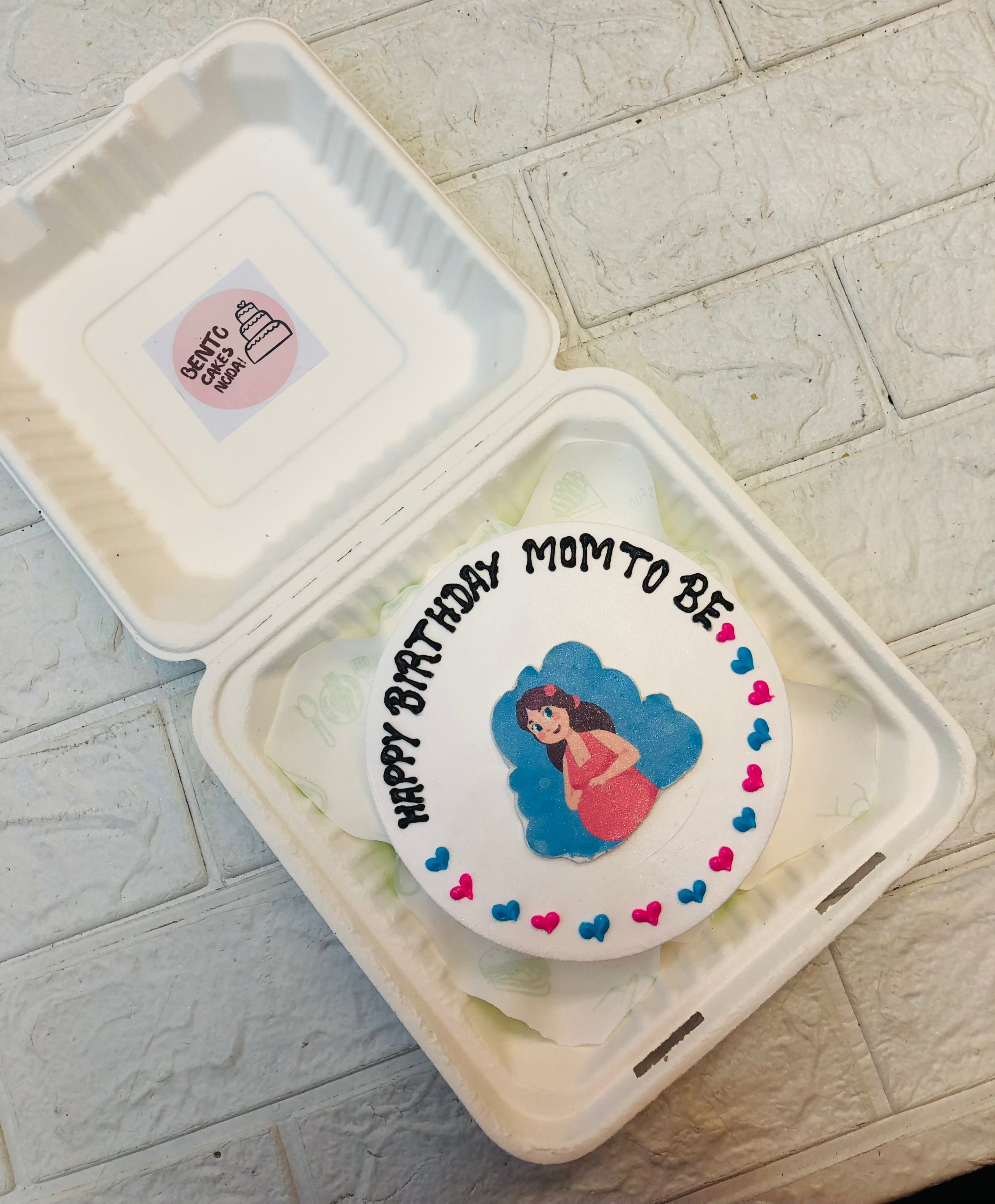 A white bento cake having Mom To Be sticker with blue and pink hearts.
