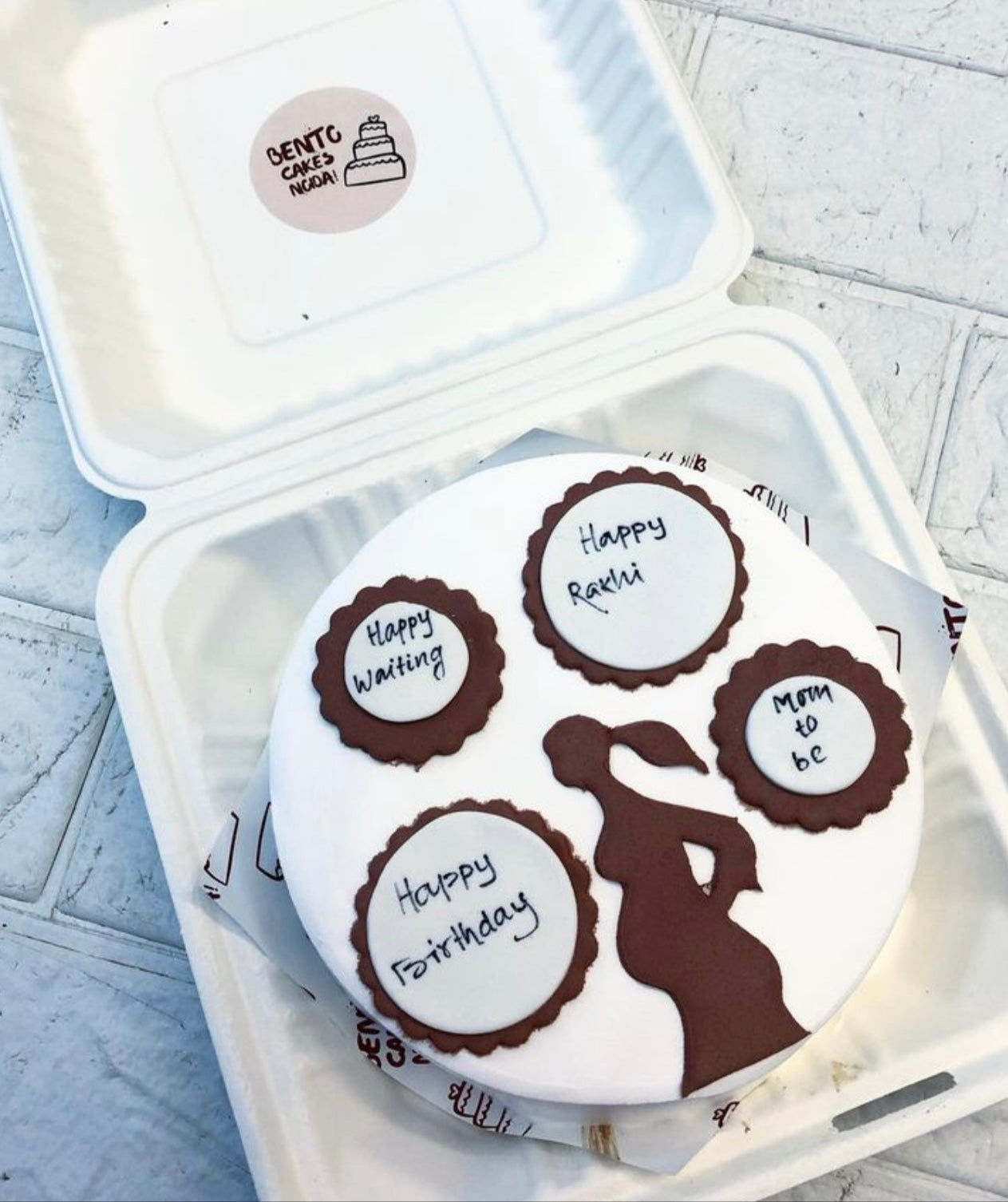 A delicately crafted white bento cake featuring chocolate decorations, including four circular plaques with messages such as "Happy Waiting," "Happy Rakhi," "Mom to be," and "Happy Birthday." The cake also has a silhouette of a pregnant woman, making it a perfect celebratory treat for moms-to-be. 