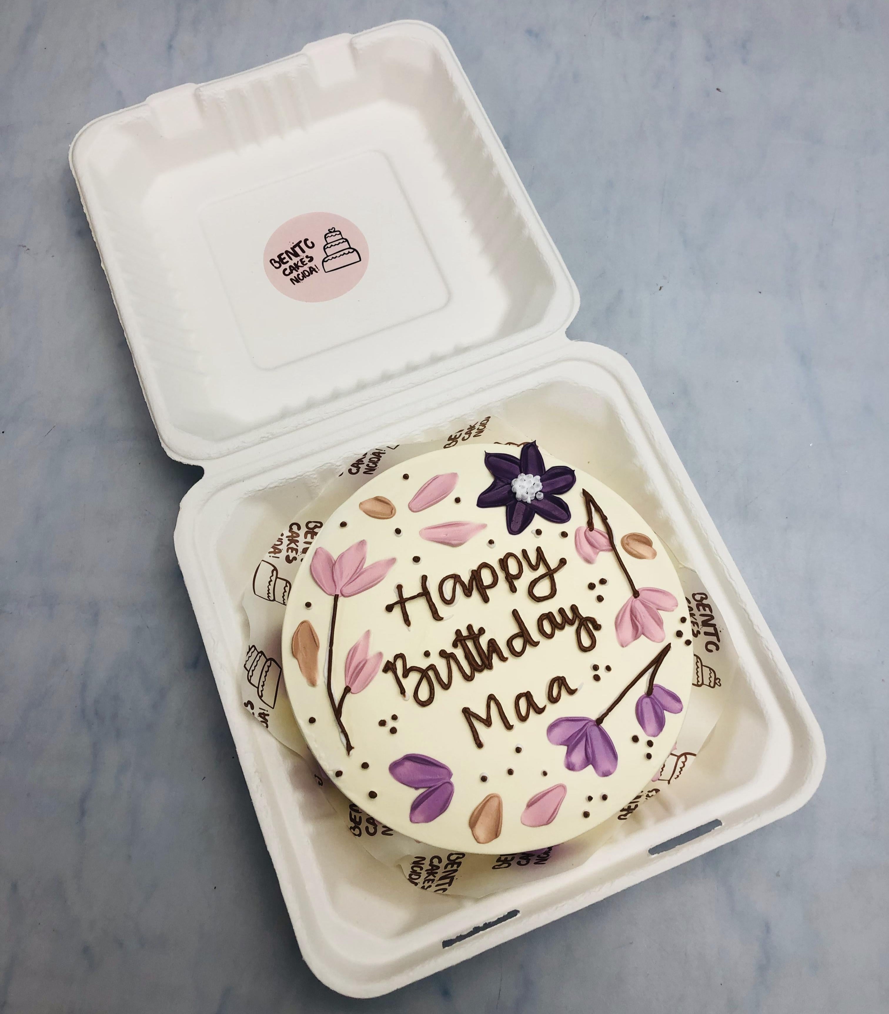A beautifully decorated birthday cake with floral designs in shades of purple and pink, placed inside a bento box. The cake has the message "Happy Birthday Maa" written on it in elegant brown icing.