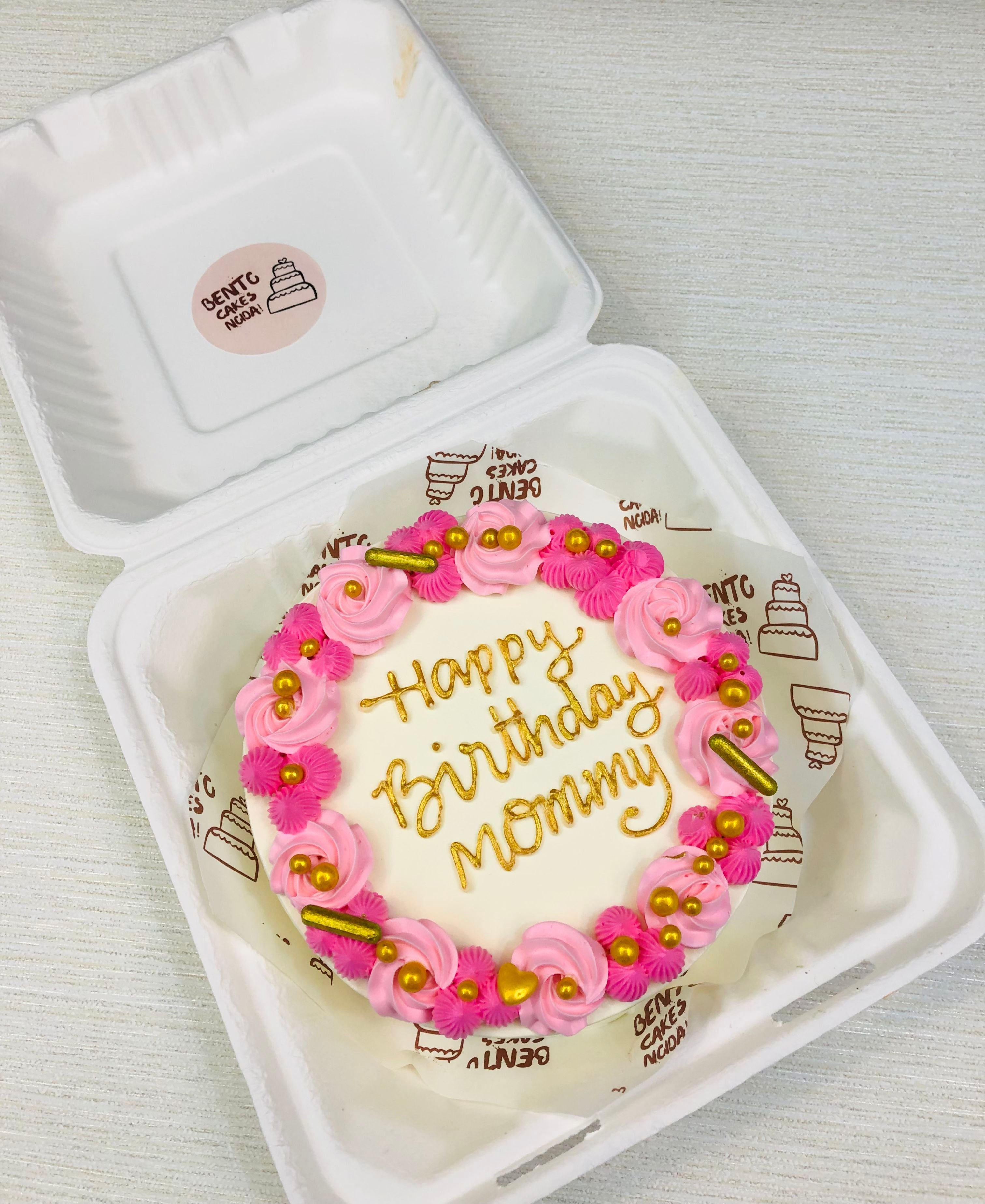 A white bento cake with pretty pink florals design on its boundary with a golde text "Happy Birthday Mommy" and the cake even decorated with golden sprinkles.