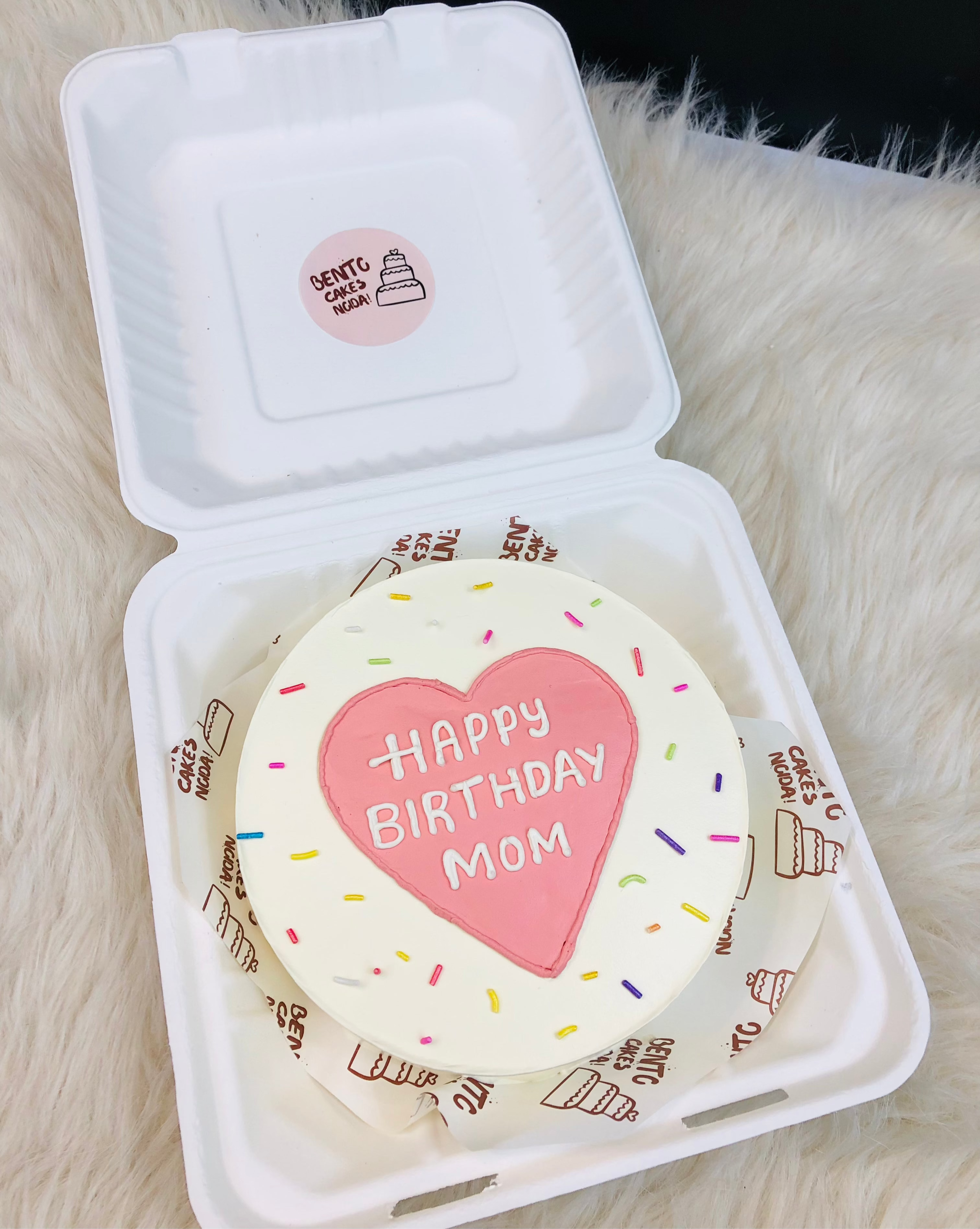A small round birthday cake with a white frosting base and colorful sprinkles, featuring a pink heart in the center with the text 'HAPPY BIRTHDAY MOM' written in white.