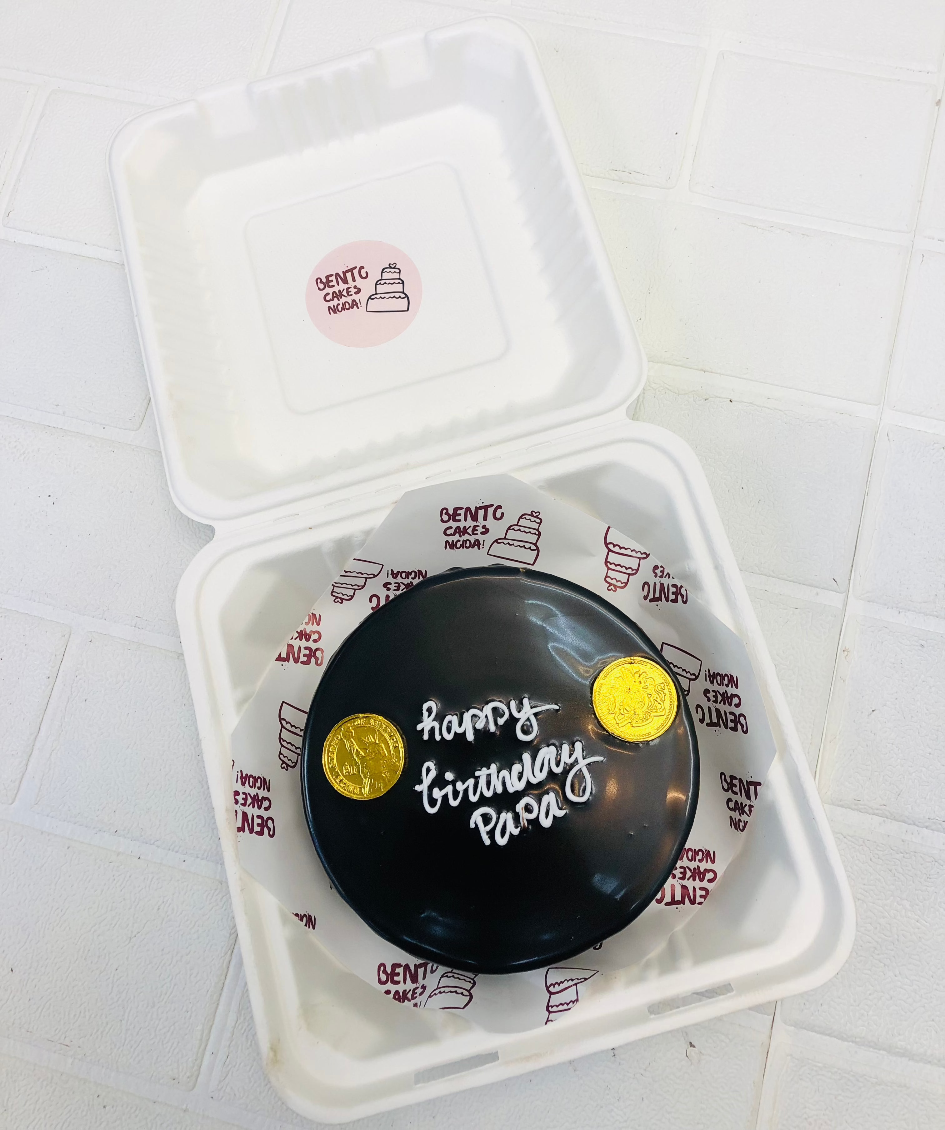 A stylish black birthday cake with "happy birthday papa" written in white icing, elegantly placed inside a bento box.