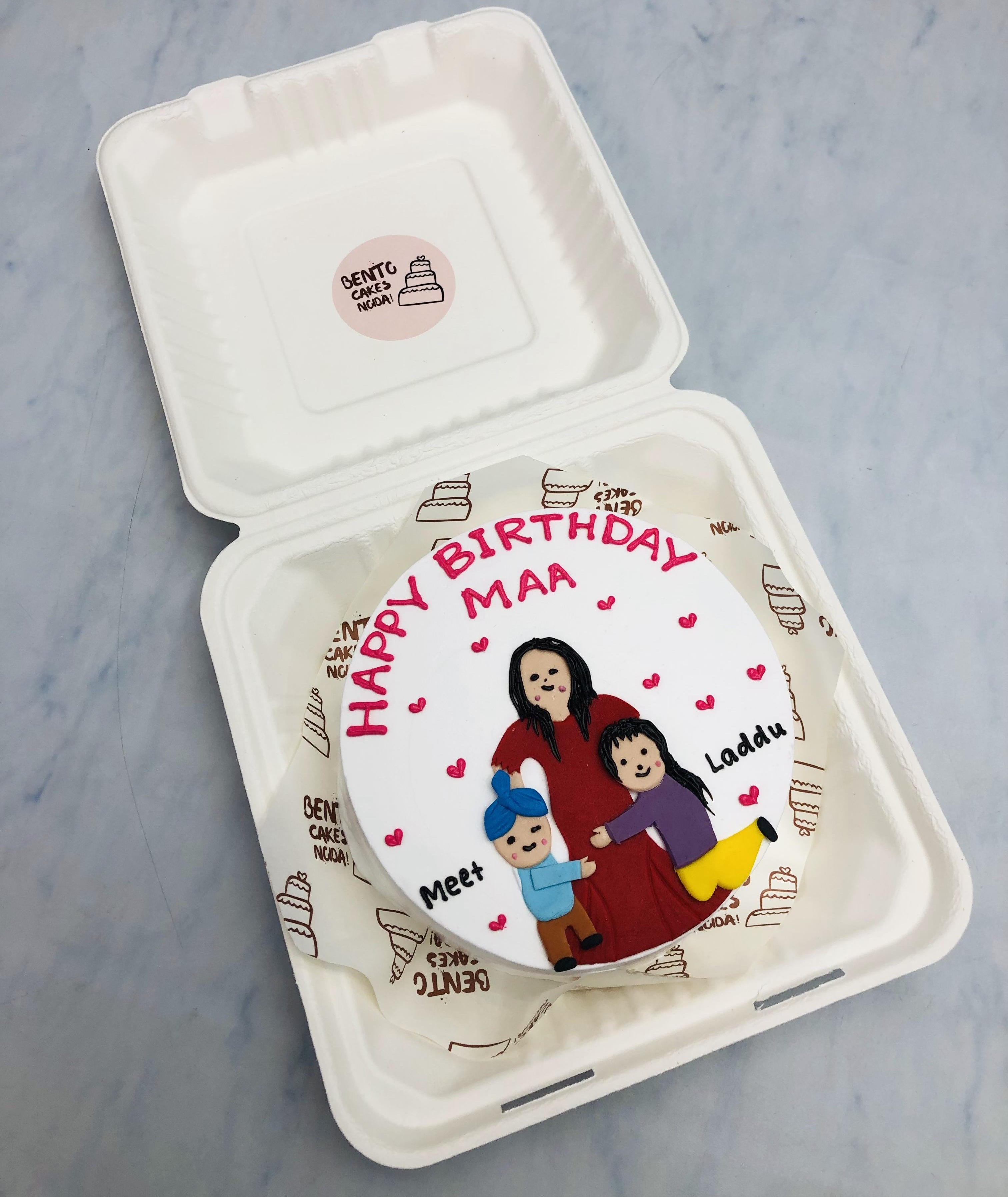 A specially crafted birthday cake for "Maa" featuring a custom illustration of two children named Meet and Laddu. The white cake is decorated with red lettering that says "Happy Birthday Maa" and a charming drawing of a woman with her children.