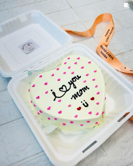 A heart shape white cake with pink and red hearts all over the cake and a beautiful text "I love you mom" written with black icing on it