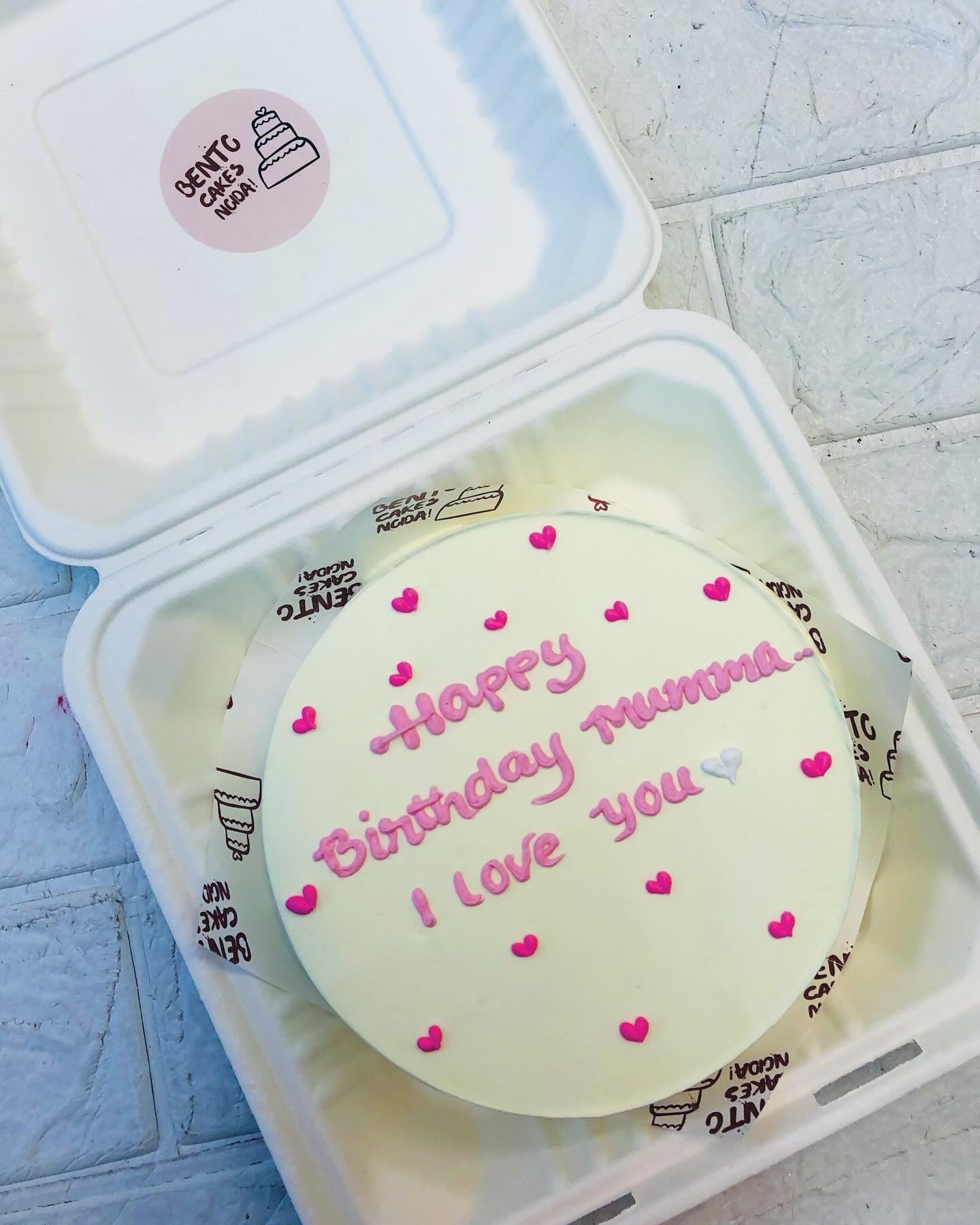 An offwhite bento cake having pink and white hearts design and a text "Happy Birthday Mumma" & "I Love You" written with light pink icing.
