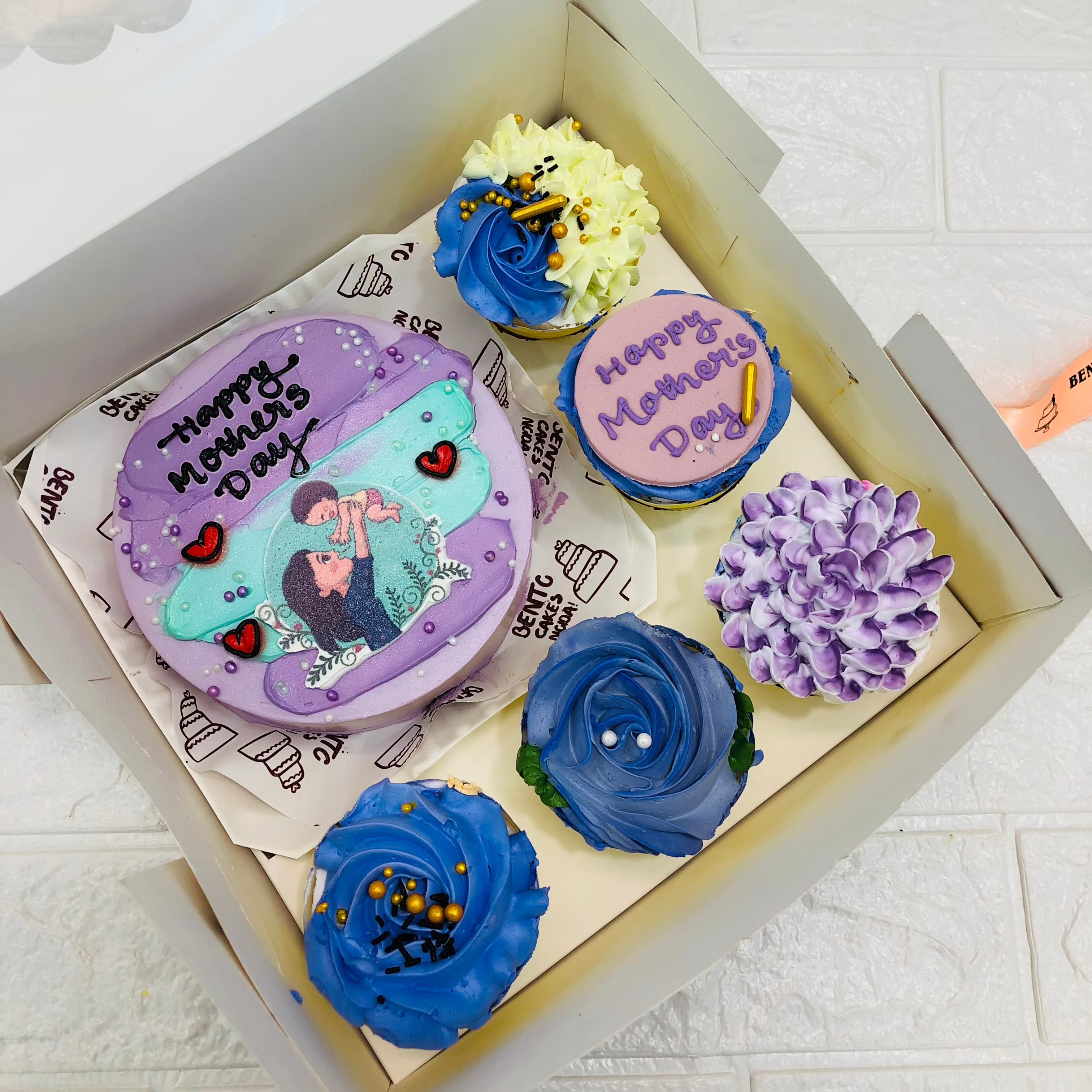 A large bento bo containing 1 cake and 5 cupcakes. An aesthetic purple cake having a mother and baby image with "Happy Mother's Day" text and cupcakes having blue and purple floral designs.