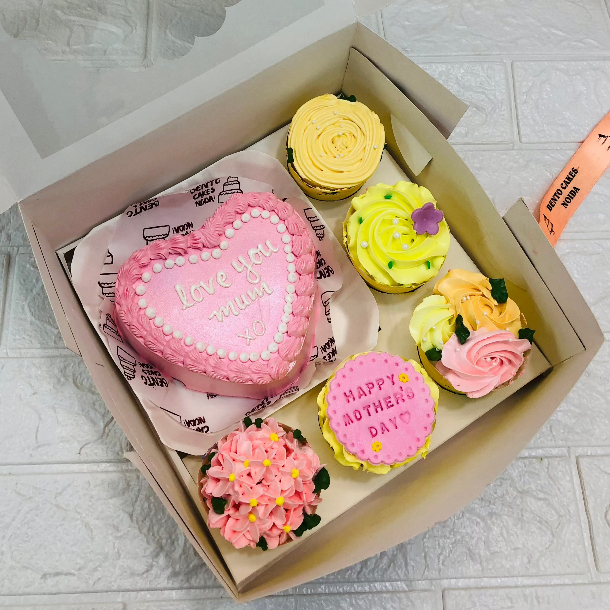 A large bento bo containing 1 cake and 5 cupcakes. A pink heart shape  cake having "love you mum XO" text and cupcakes having yellow and pink floral designs.