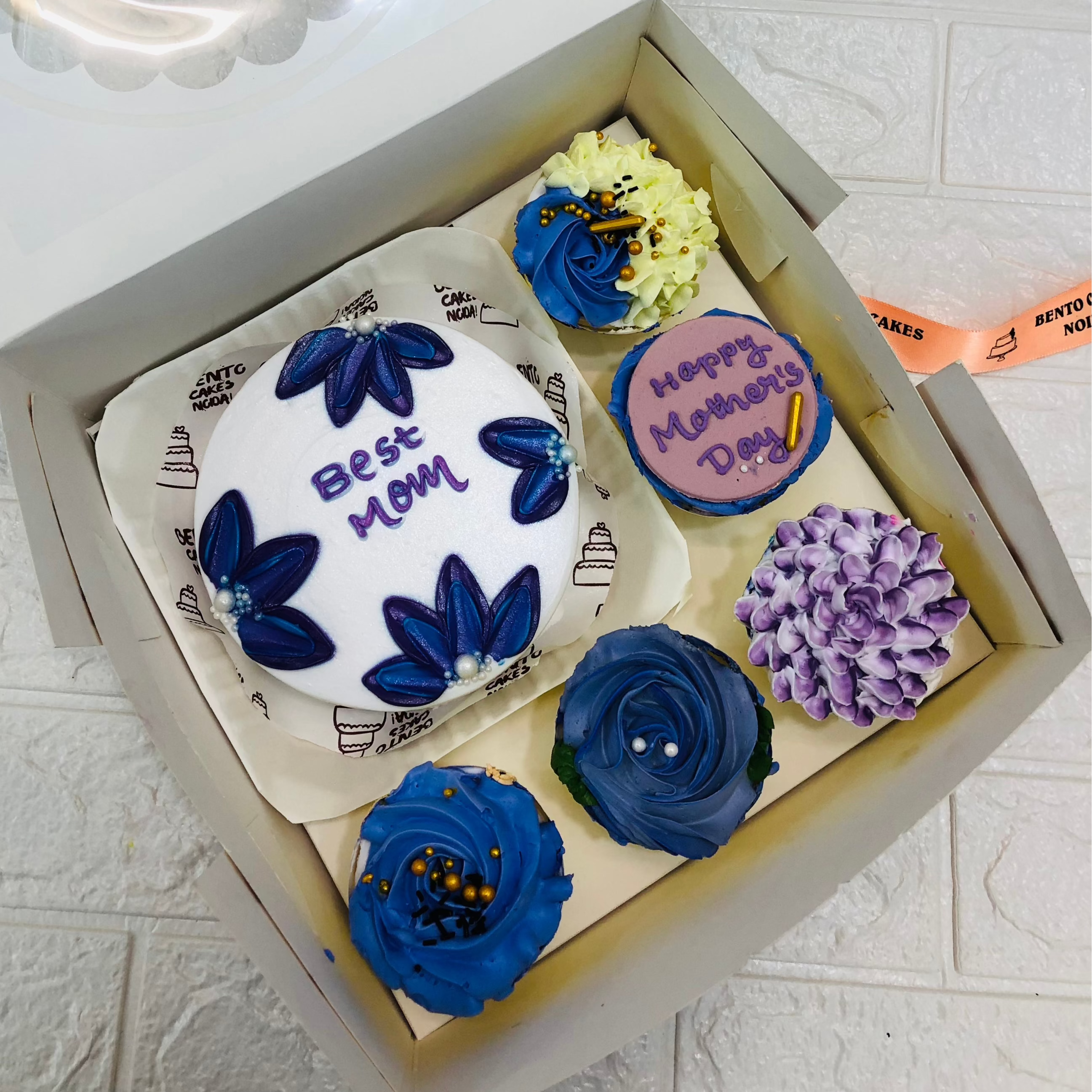 A large bento bo containing 1 cake and 5 cupcakes. A blue floral design cake having "Best Mom" text and cupcakes having blue and purple floral designs.