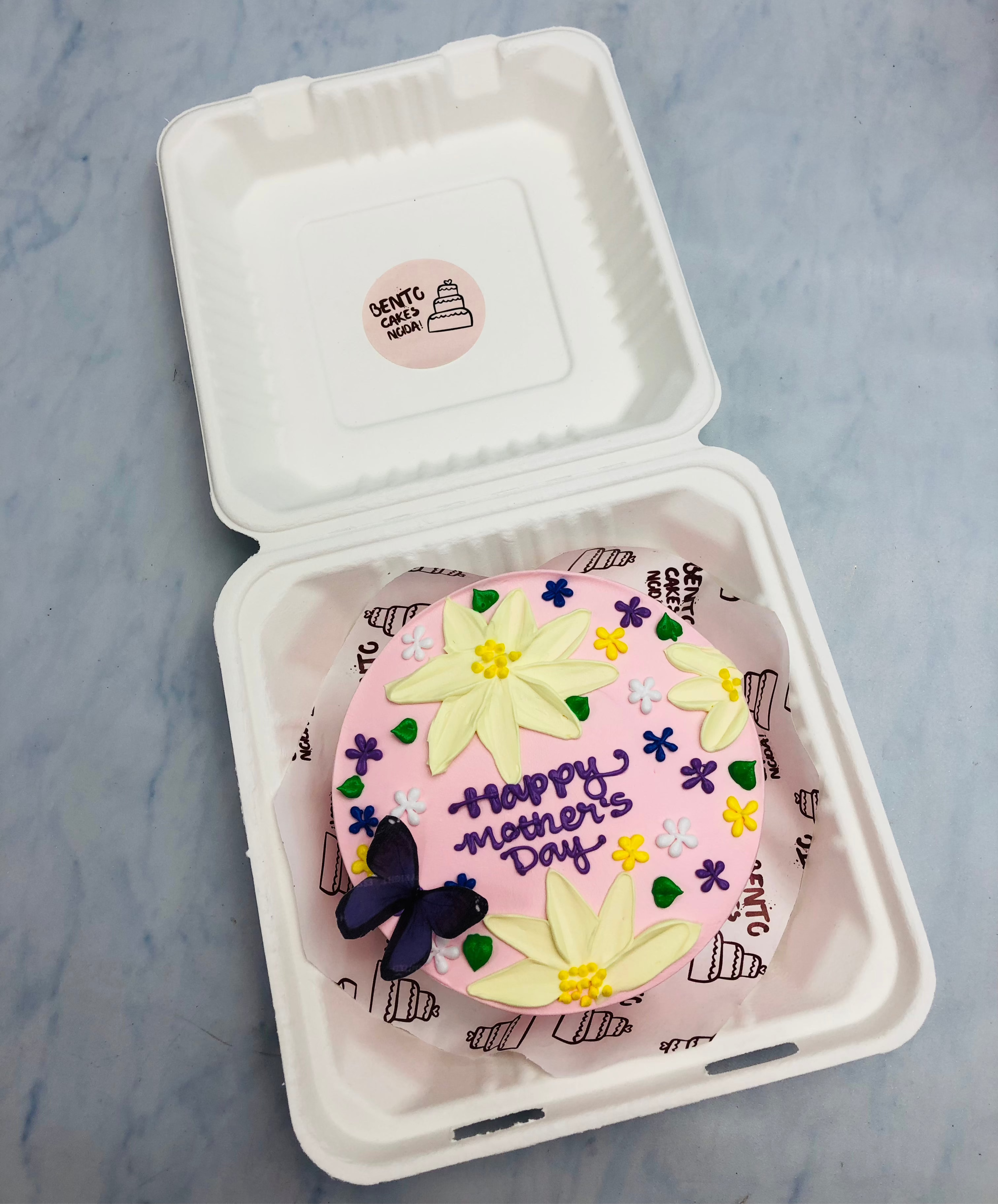 A light pink bento cake having yellow flowers design with a blue butterfly and is designed for mother's day occasion.