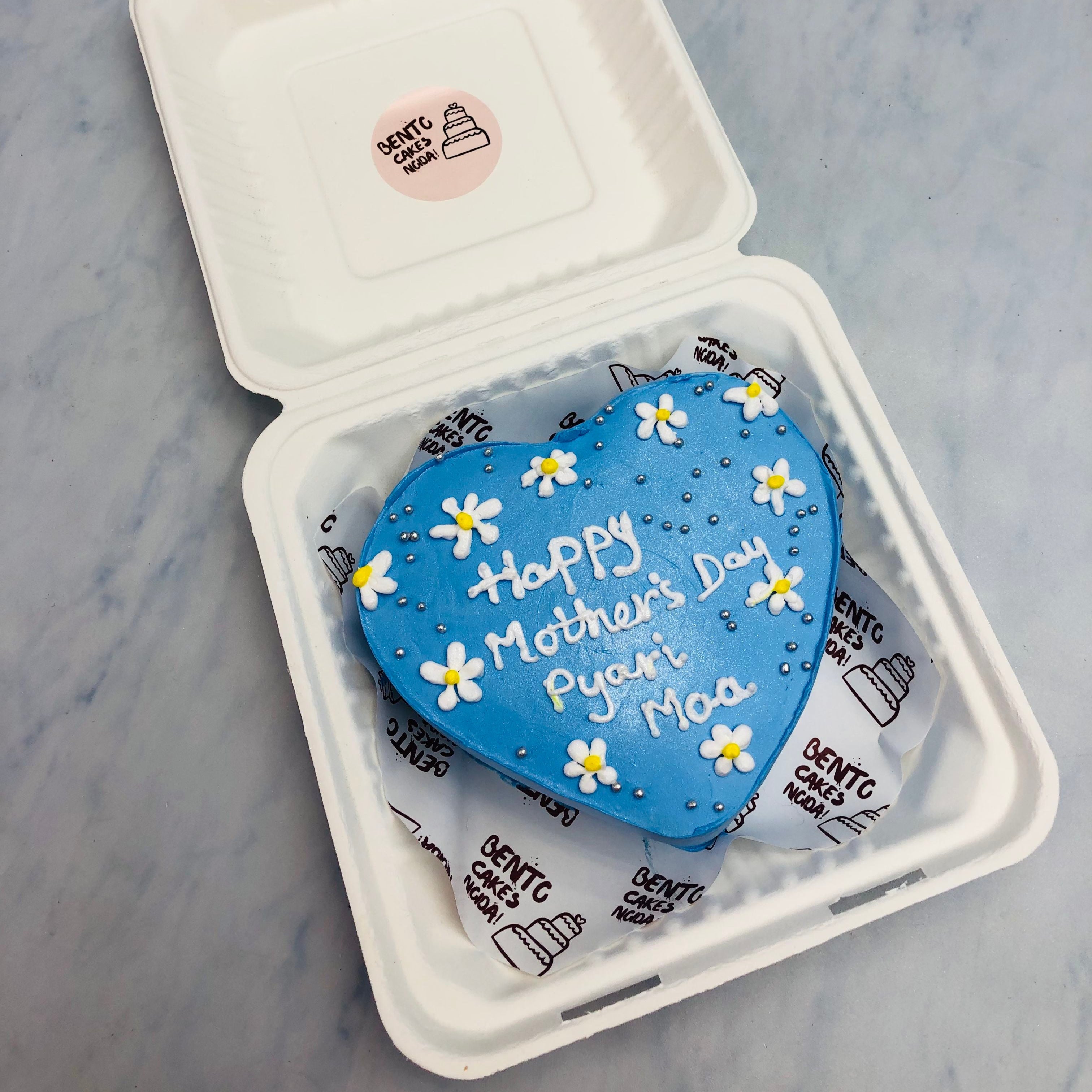 A blue heart shape cake decorated with pretty flowers and is designed for Mothers Day Occasion.
