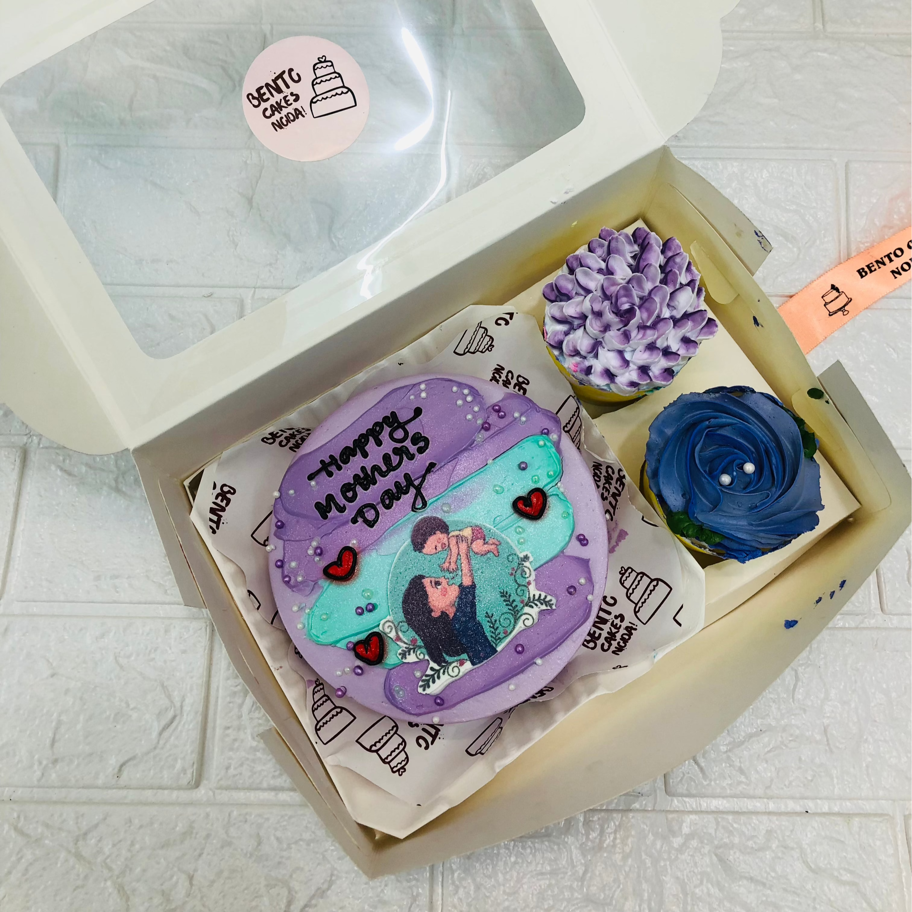 A mini bento combo having 1 cake and 2 cupcakes set. A purple color  cake have aesthetic deign with mother and child print on it and cupcakes having dark blue and purple floral design.