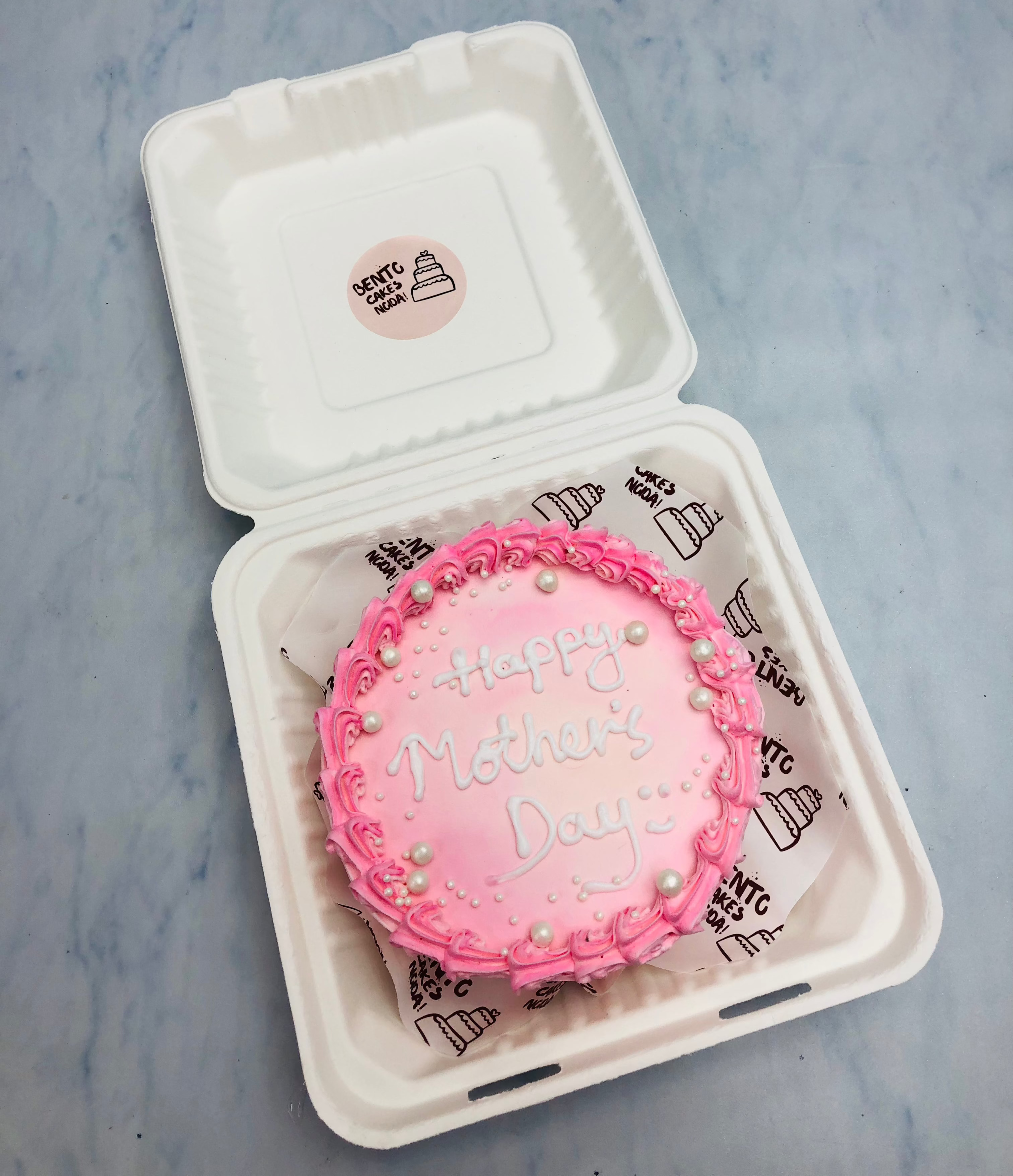 A shiny pink bento cake with floral design on its boundary and decorated  with white sprinkles for mother's dat.