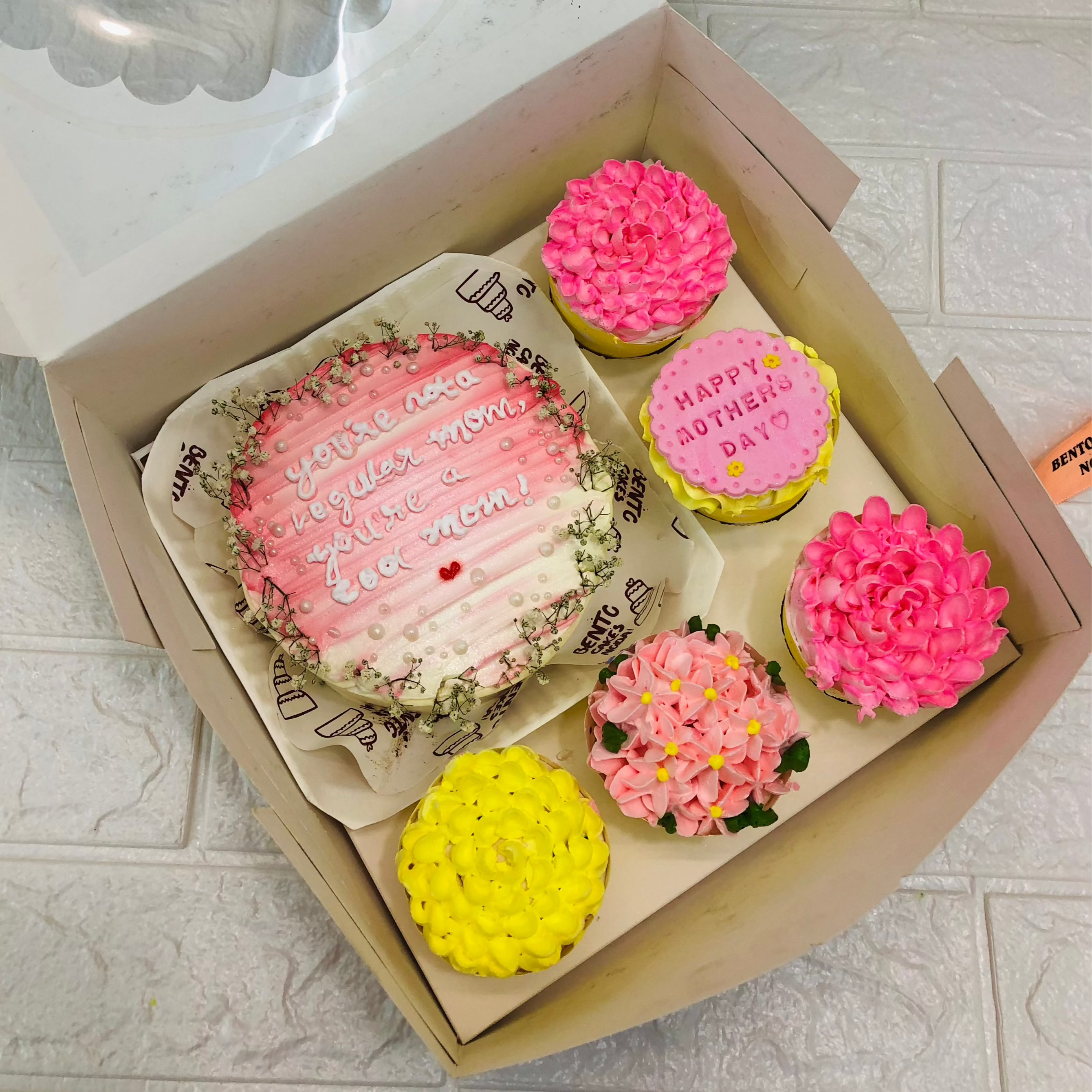 A large bento bo containing 1 cake and 5 cupcakes. An aesthetic pink cake having a text and cupcakes having pink and yellow floral designs.