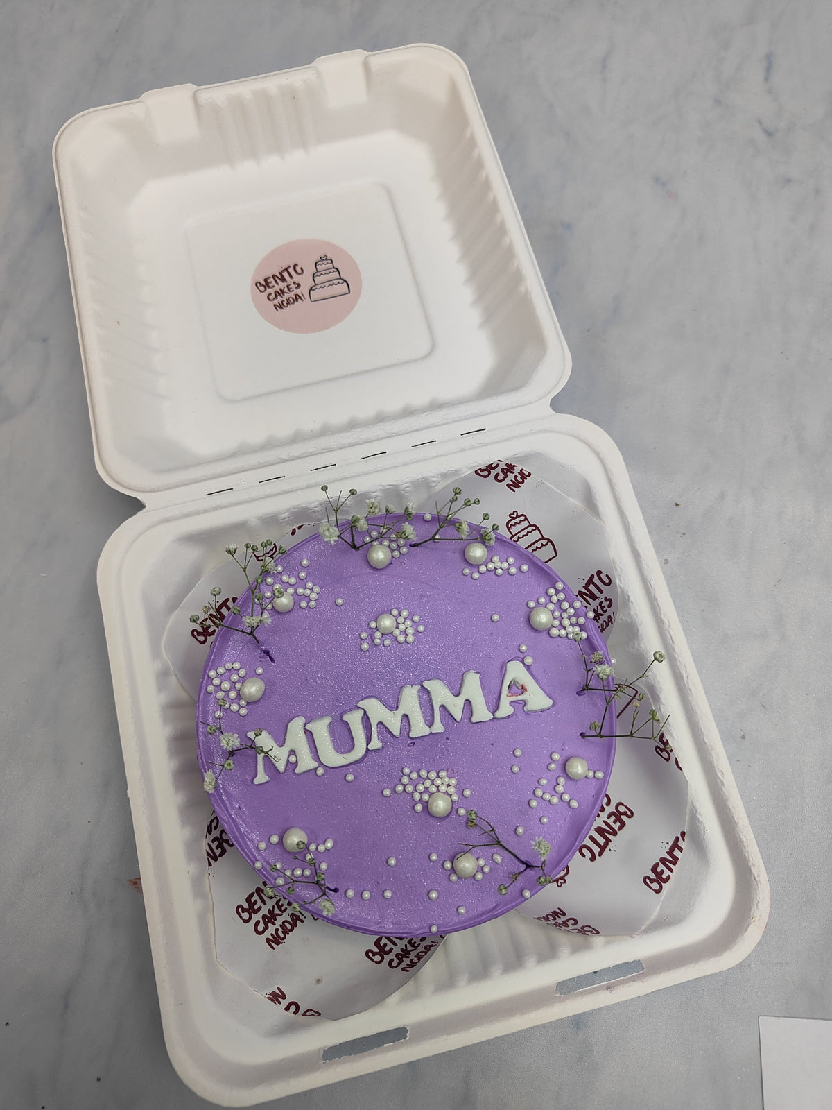 An aesthetic purple bento cake with fondant text Mumma written and decorated with floral and white spinkles.