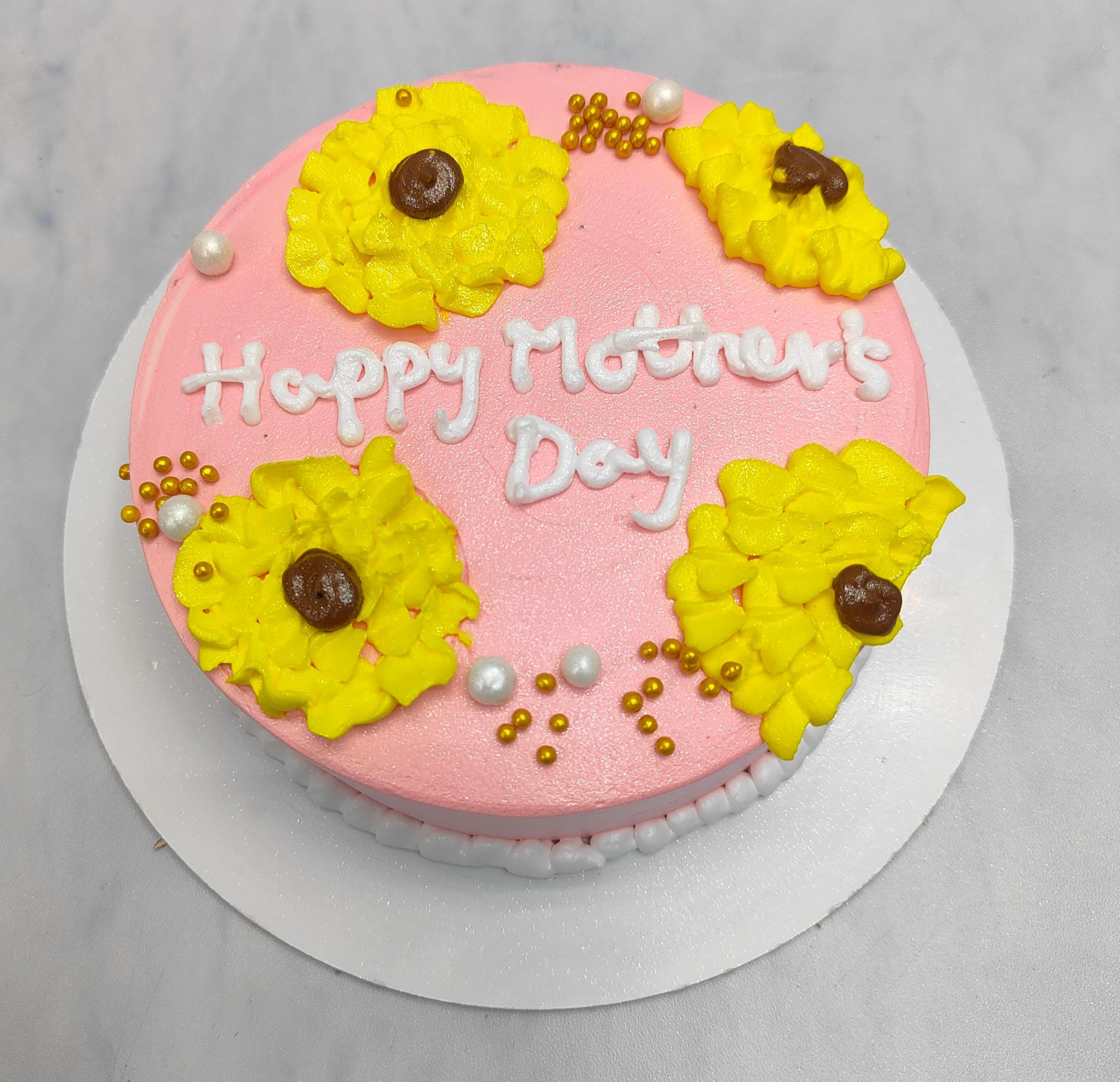 A light pink icing cake with yellow floral design on it and a "Happy Mother's Day written with white icing".