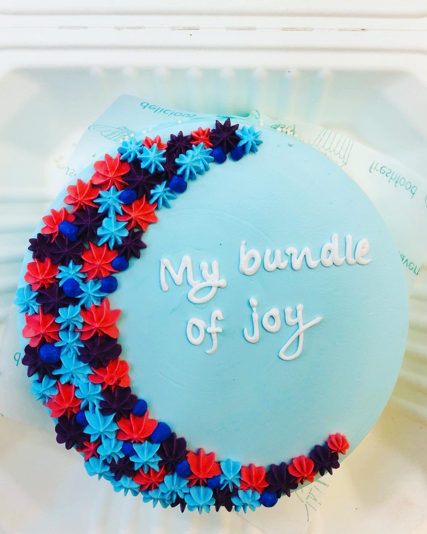 A light blue cake decorated with colorful florals design on one side of round cake and a text "My bundle of joy" written with white icing on it. 