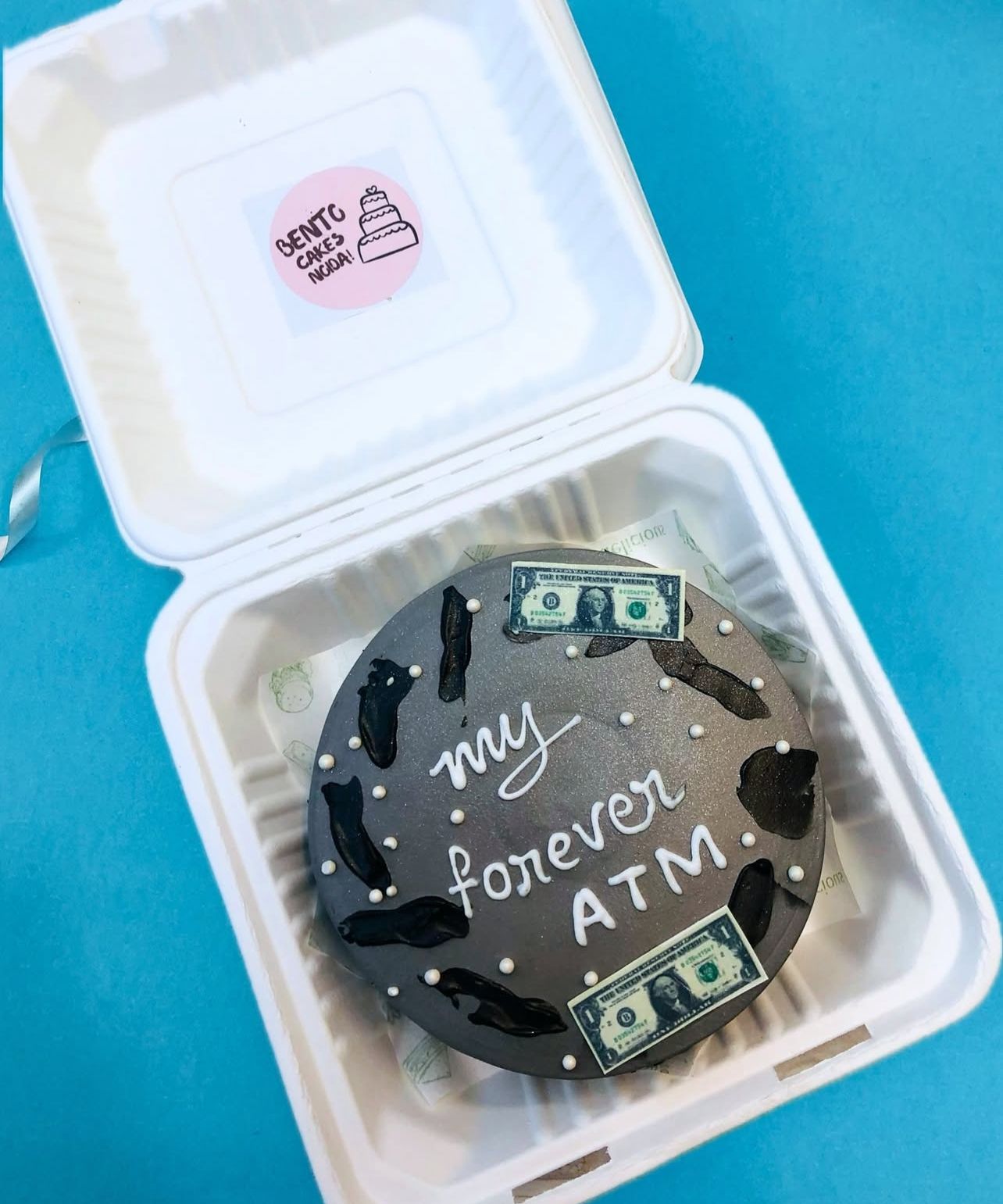 A dark grey cake with the phrase 'my forever ATM' written in white icing, decorated with edible dollar bills and black shapes inside a bento box.