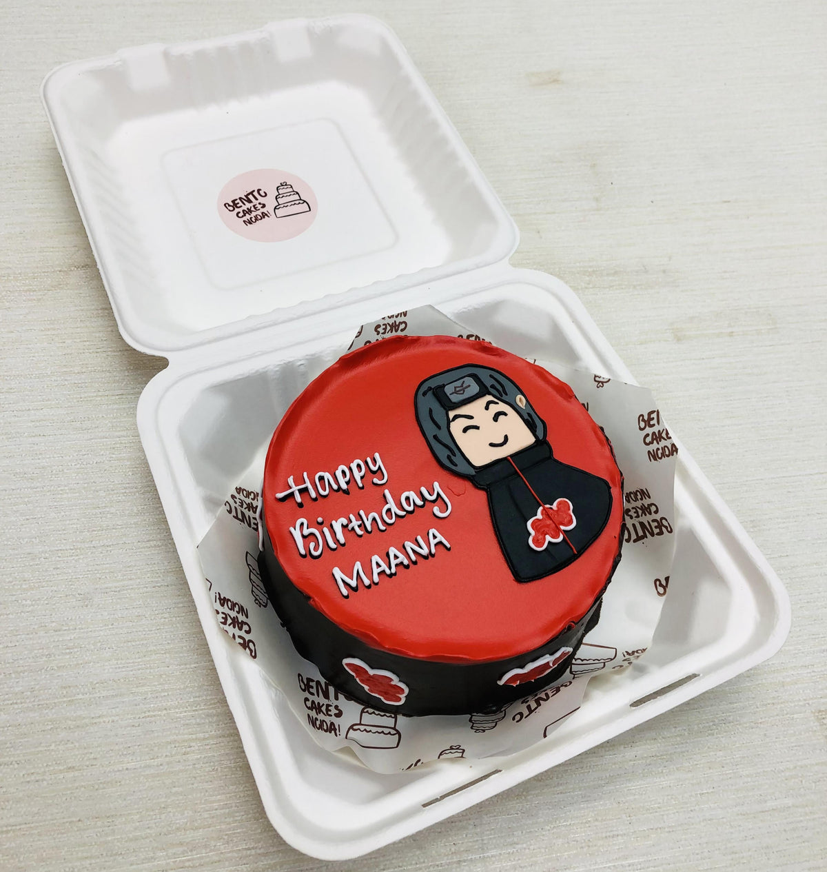 A small round birthday cake with a red top and black sides, featuring a hand-drawn character from Naruto and the text 'Happy Birthday MAANA'. 