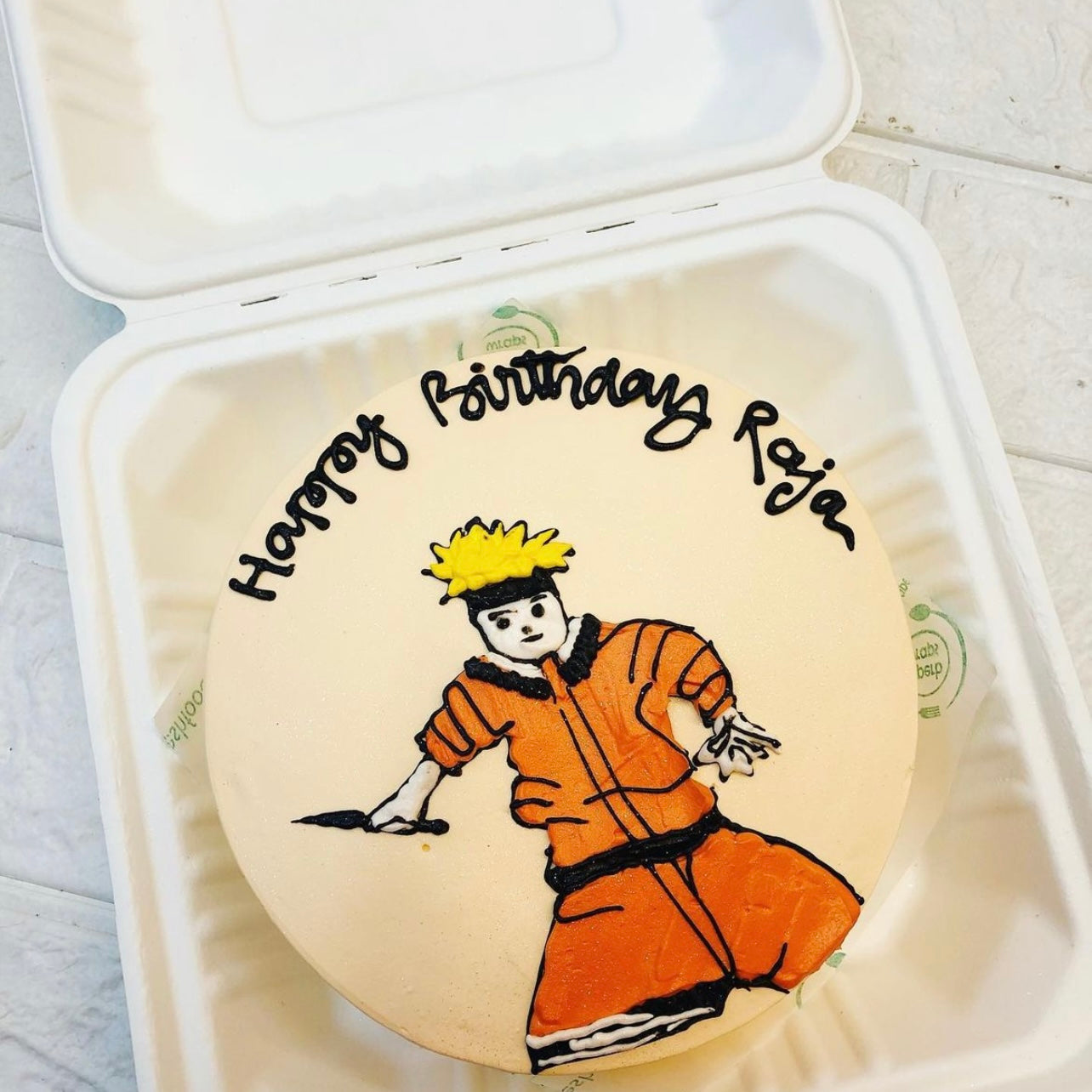 A round birthday cake with a Naruto-themed design, featuring an illustration of Naruto Uzumaki in his iconic orange outfit. The cake is placed inside a bento box. The text "Happy Birthday Raja" is written in black icing around the character.