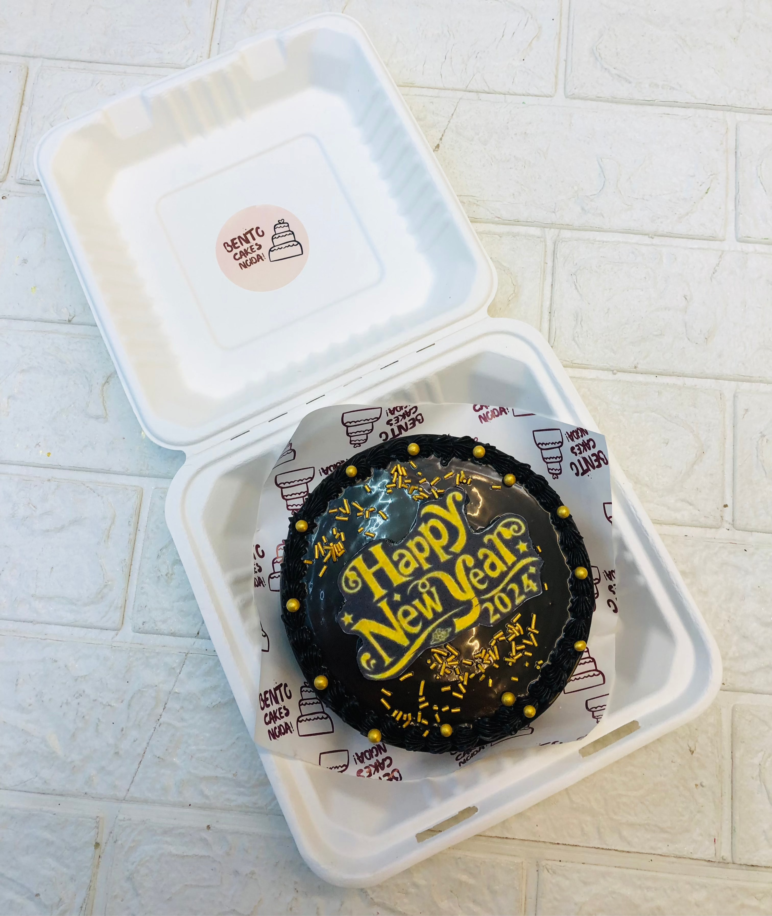 A black chocolate bento cake decorated with golden sprinkles and a happy new year print placed on it.