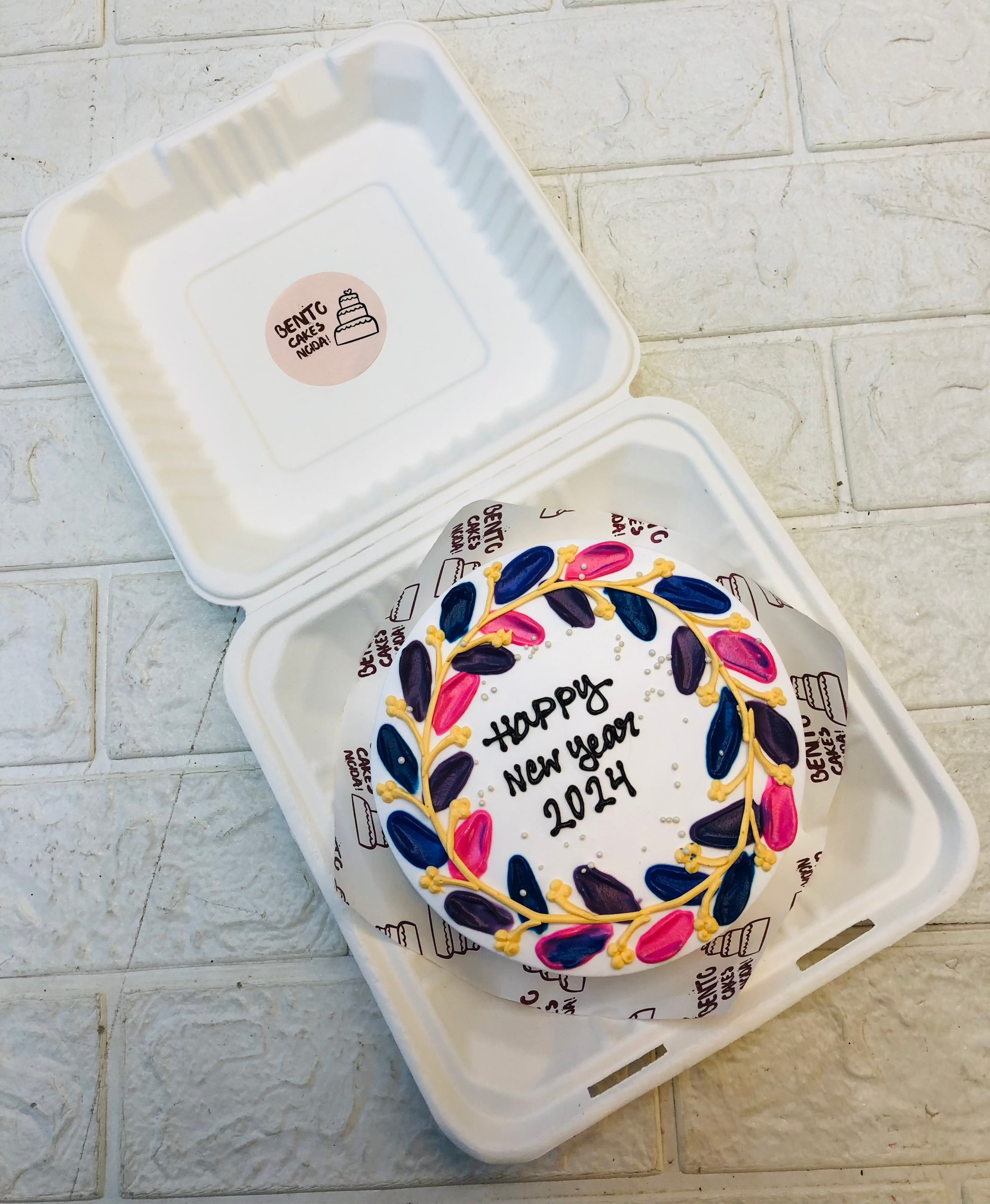 A white bento cake having colorful floral pattels design on its border with a Happy New Year text in the center.