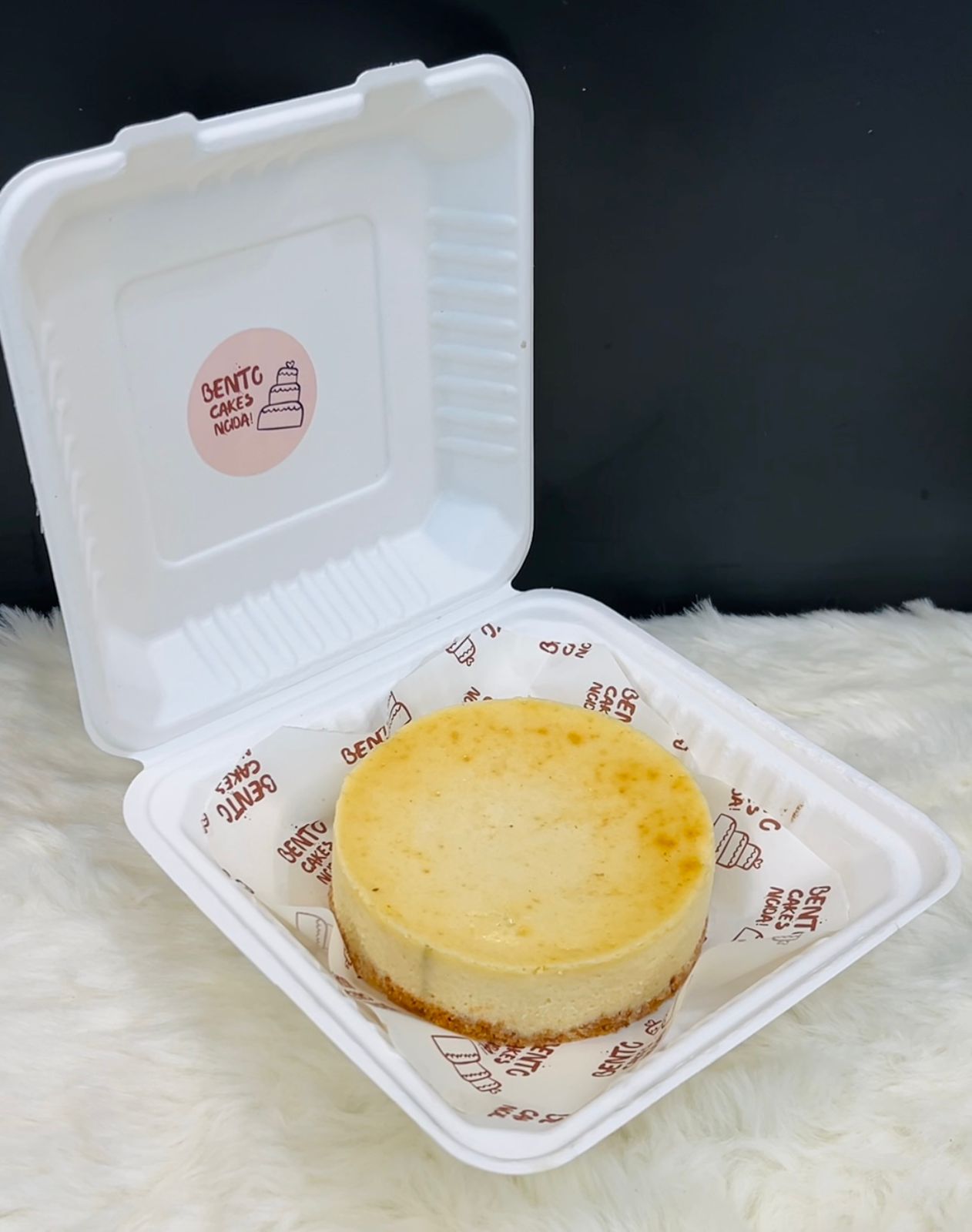 A New York cheesecake in a sleek white bento box, placed on branded parchment paper with ‘BENTO CAKES NOGA’ printed, showcasing its creamy texture and golden crust on a soft white surface.