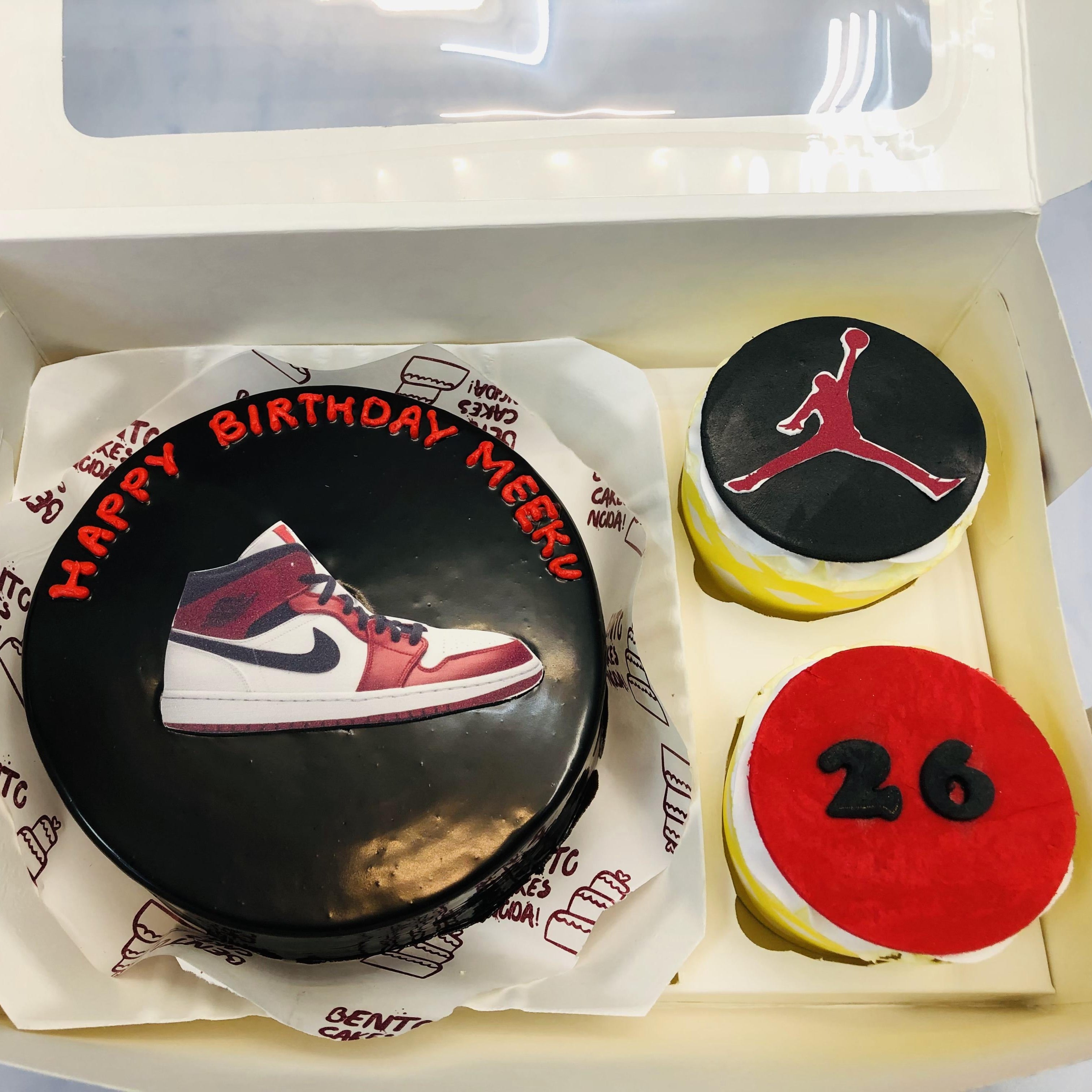 A black birthday cake with a red and white sneaker design, accompanied by two cupcakes: one with a red "26" design and another with a black Jumpman logo. The set is presented in a white box.